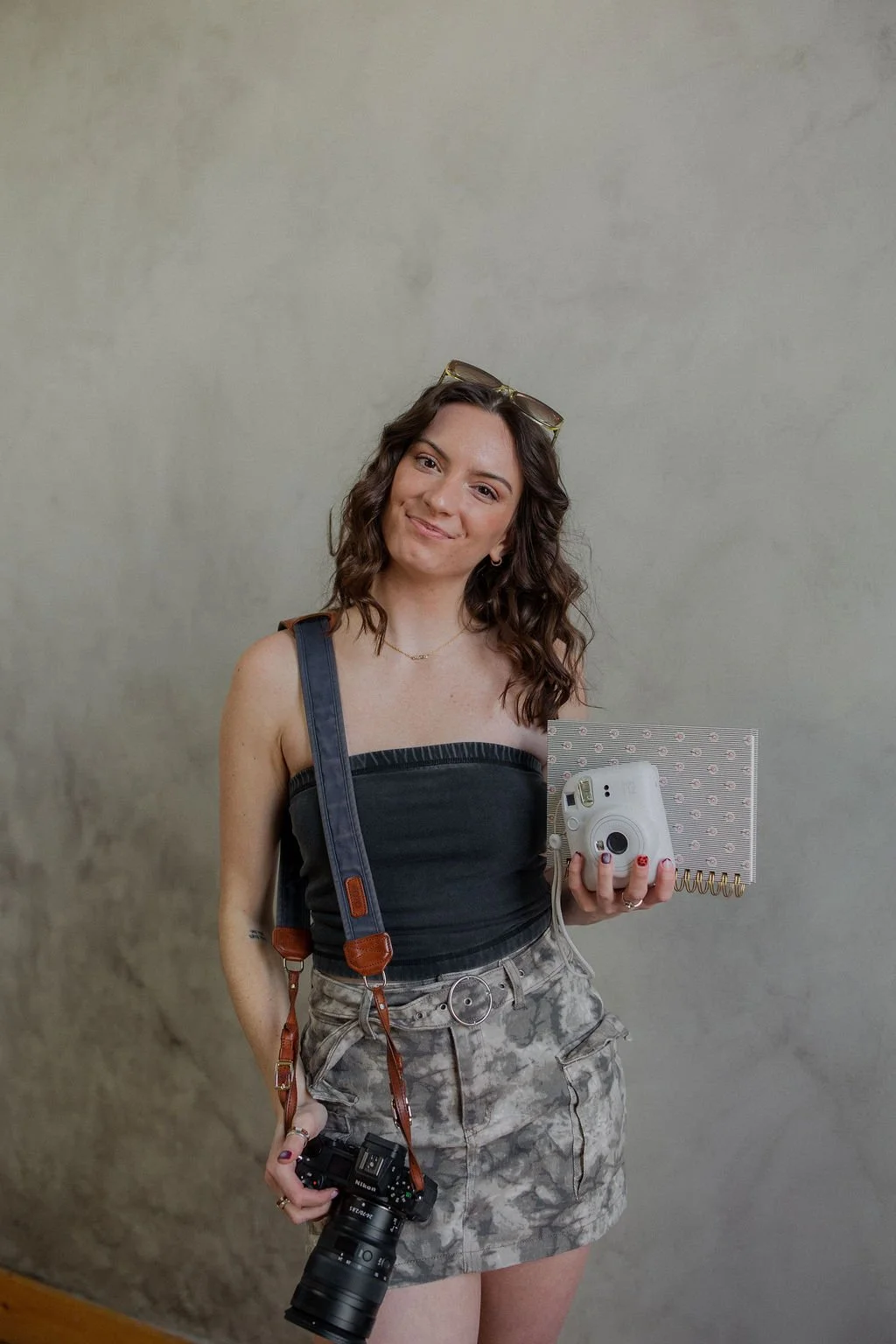 Young woman with wavy brown hair taking a mirror selfie, holding a camera in her left hand and a polaroid camera and spiral notebook in her right hand, wearing sunglasses on her head, a black strap top, and patterned shorts.