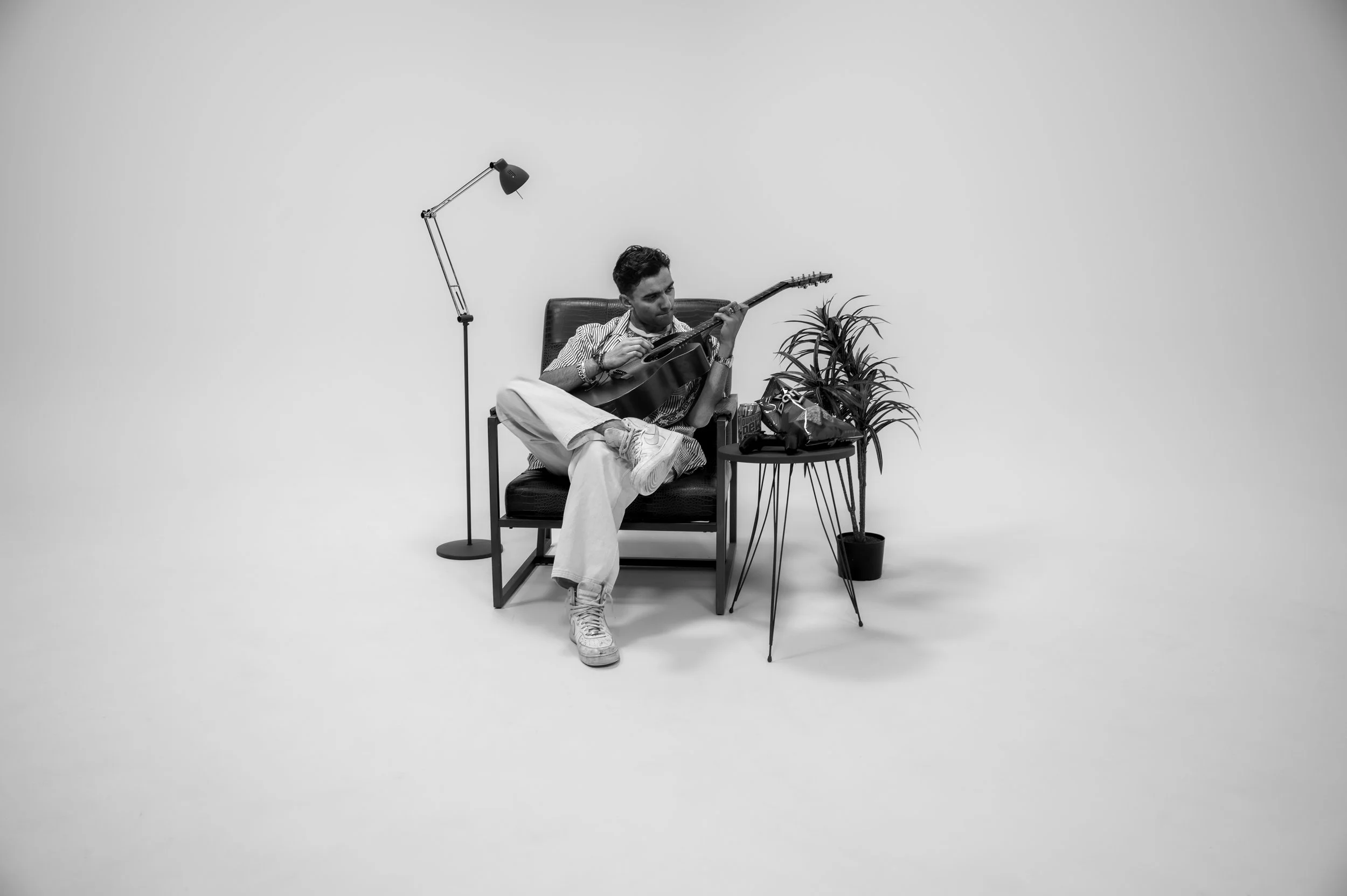 A young man sitting in a chair in a minimalistic room, playing an acoustic guitar. Beside him is a small table with snacks, a drink, and a potted plant. A tall floor lamp is positioned behind him. The room has plain white walls and no other furniture