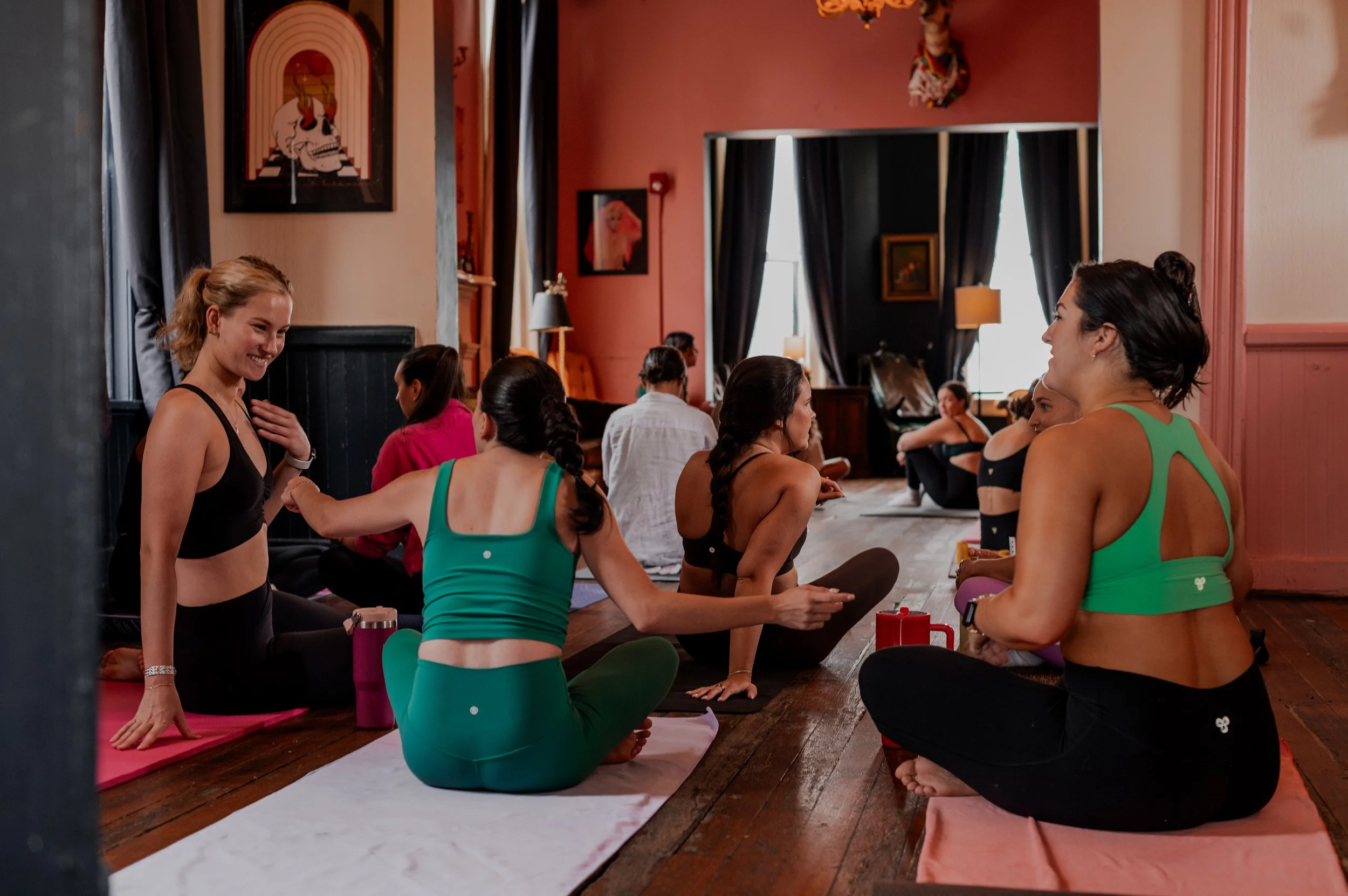 Group of women practicing yoga and meditation in a cozy room with pink and black walls, framed artwork, and large windows with dark curtains.