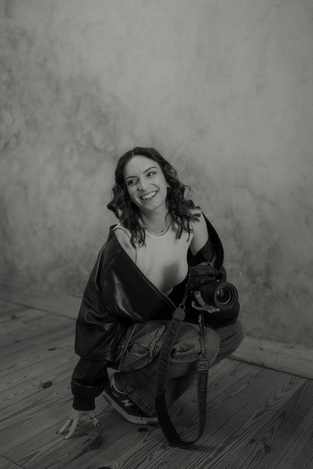 A young woman with curly hair smiling while holding a camera, squatting on a wooden floor against a textured wall.