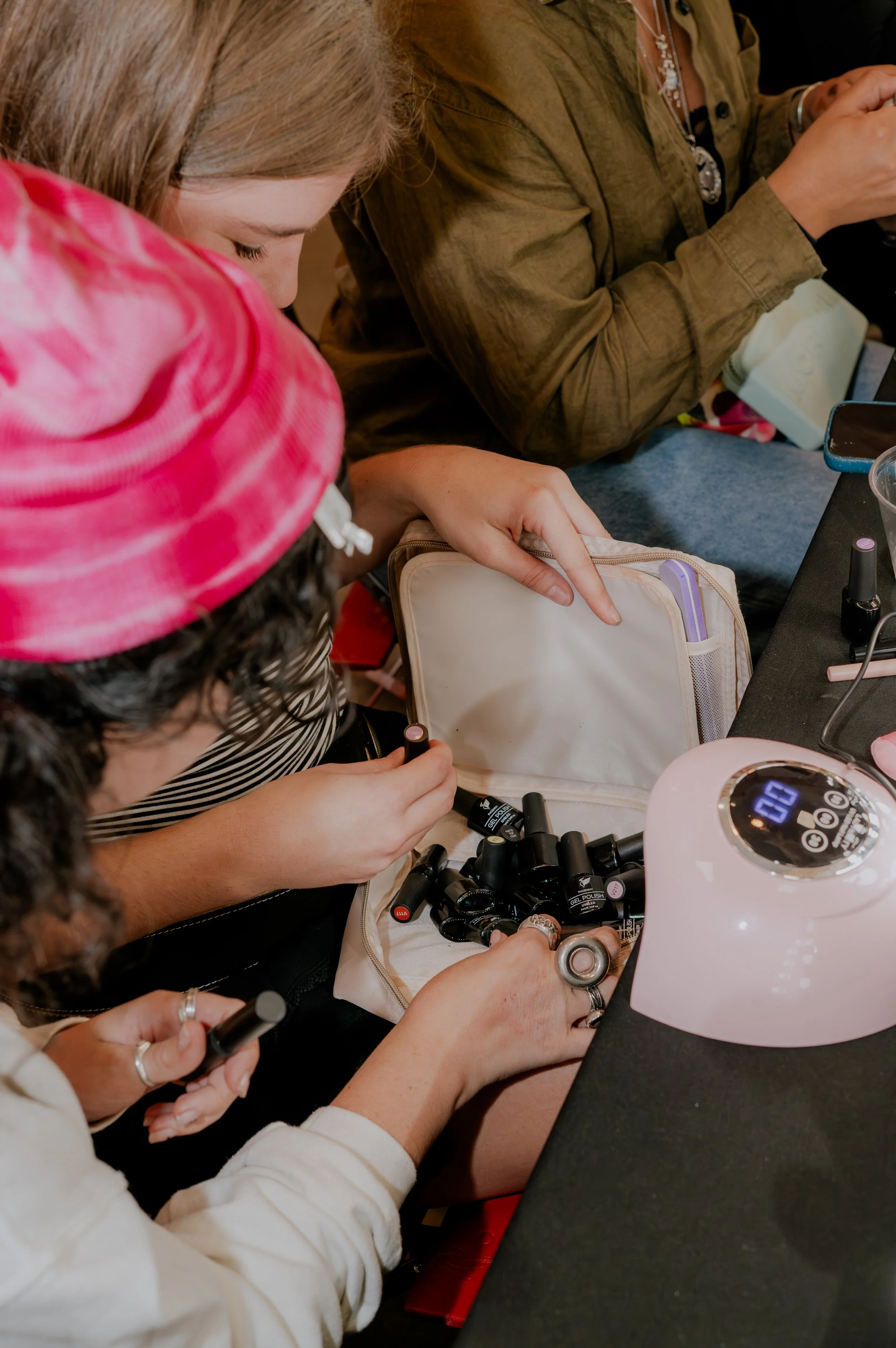 A woman and a young girl sitting at a table, applying black gel nail polish to their nails. The table has nail polish bottles, a white LED nail dryer, and a small zippered pouch. The woman is wearing a striped top and a ring, while the girl is wearin
