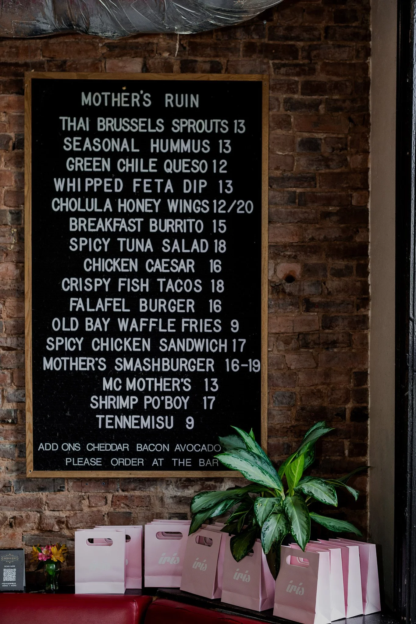 A blackboard menu in a restaurant listing various food items and prices, with a brick wall background, green plant, and pink takeaway bags on a table below.