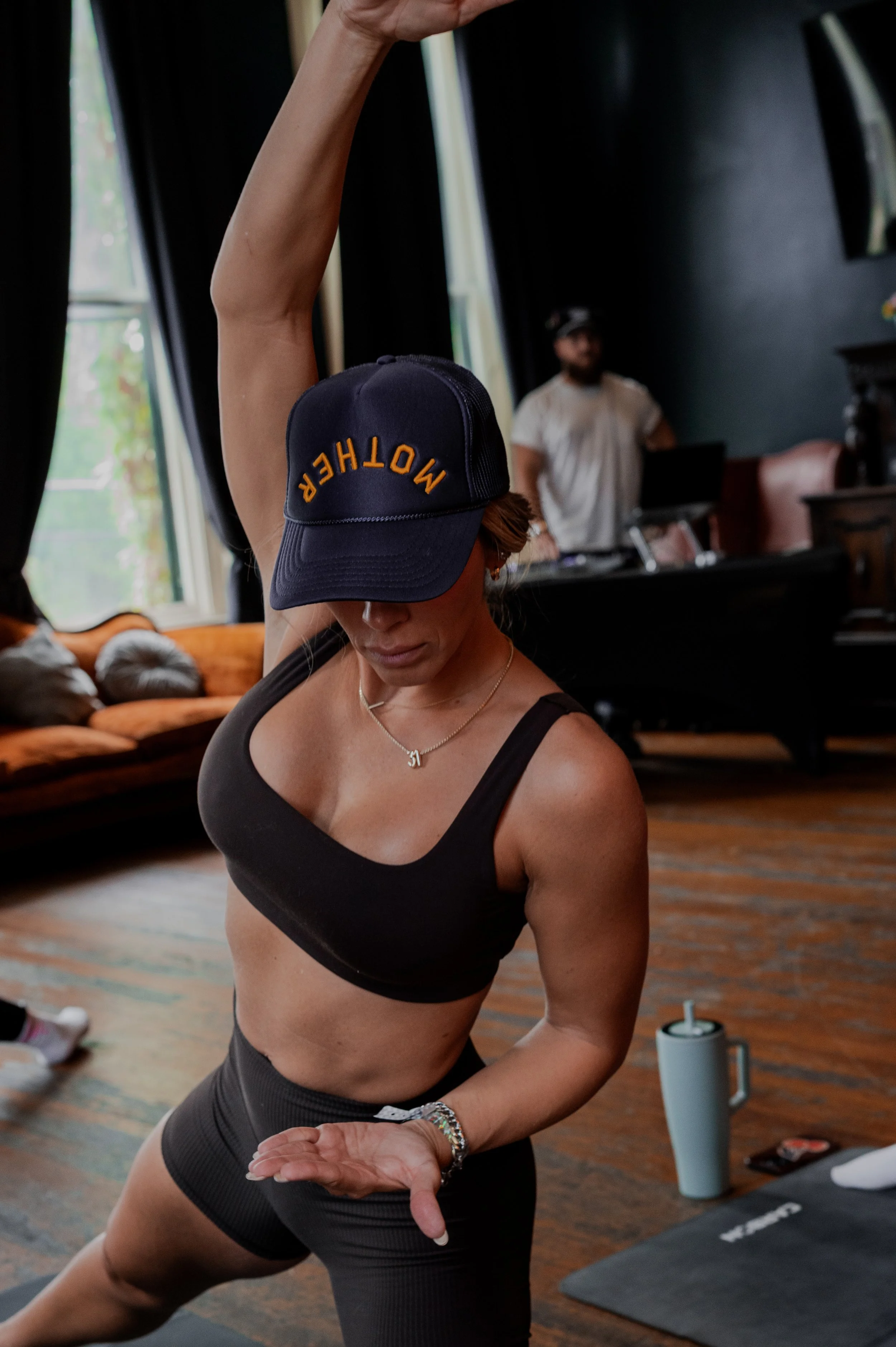 A woman in workout clothes and a 'MOTHER' cap is practicing yoga in a studio, with a DJ or instructor in the background.