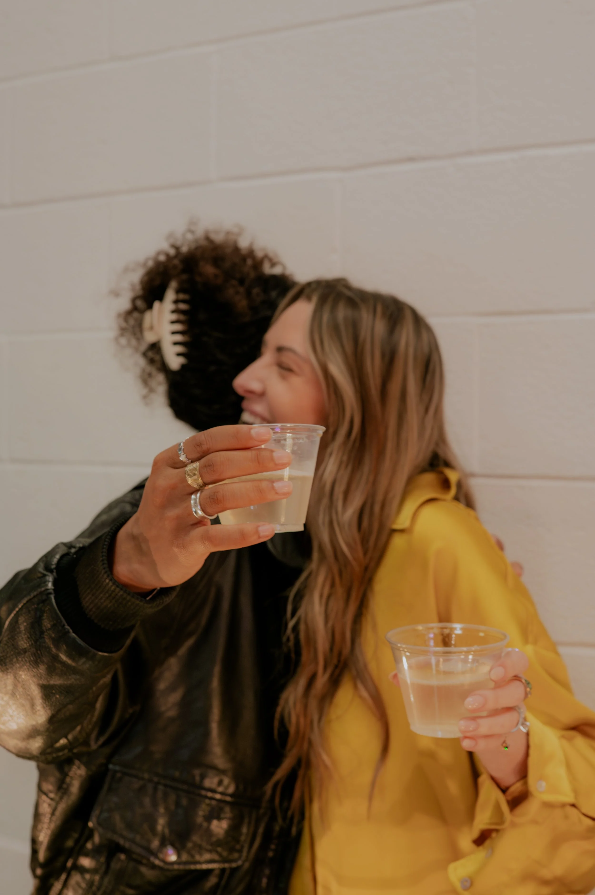 Two women sitting together, one wearing a yellow jacket and the other wearing a black leather jacket, each holding a plastic cup, with the woman in yellow smiling and the woman in black using a hairbrush in her hair.