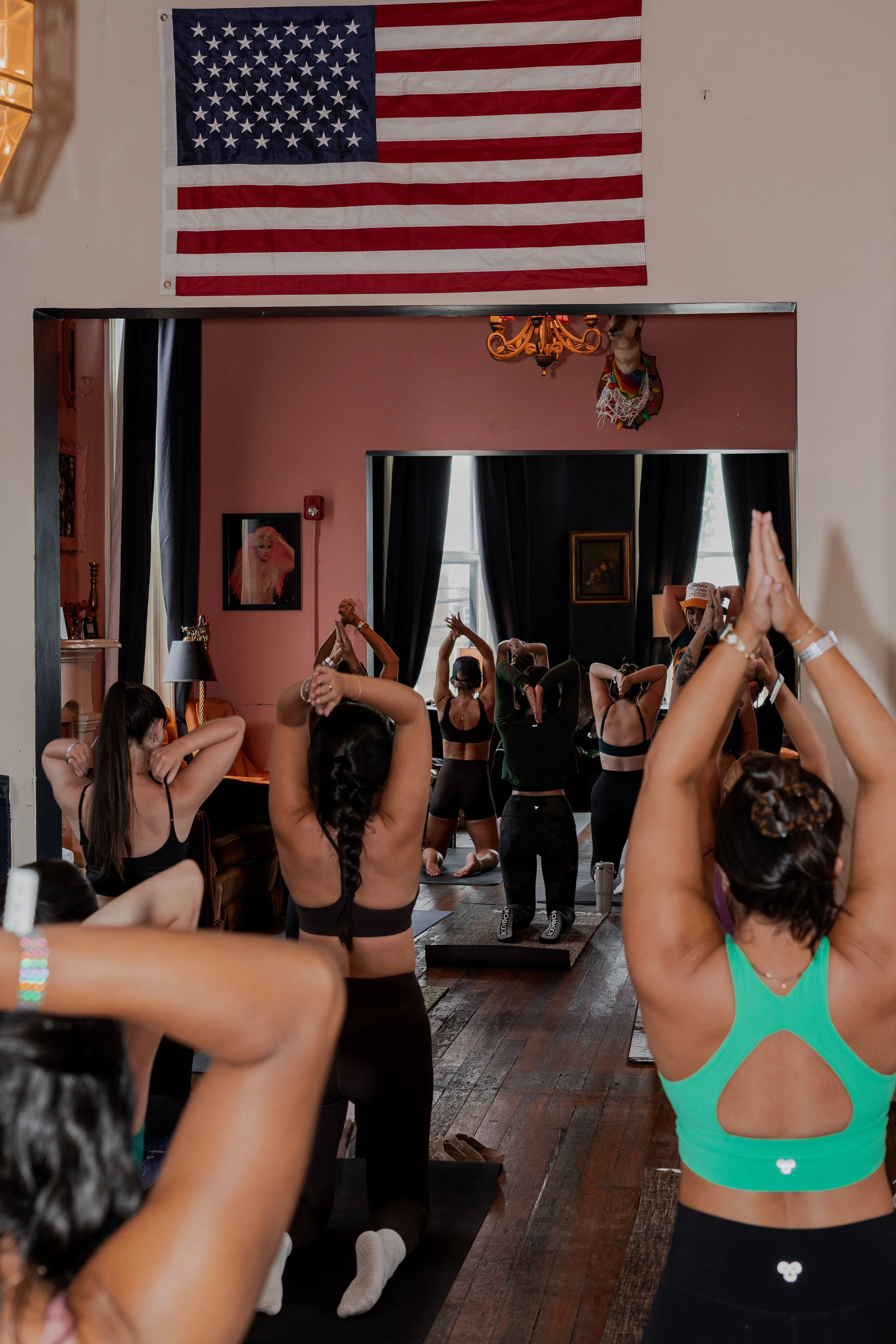 A group of people participating in a yoga class in a room decorated with an American flag and photos on the walls. The participants are stretching or doing yoga poses, some with their hands above their heads, inside a cozy, artistically decorated spa