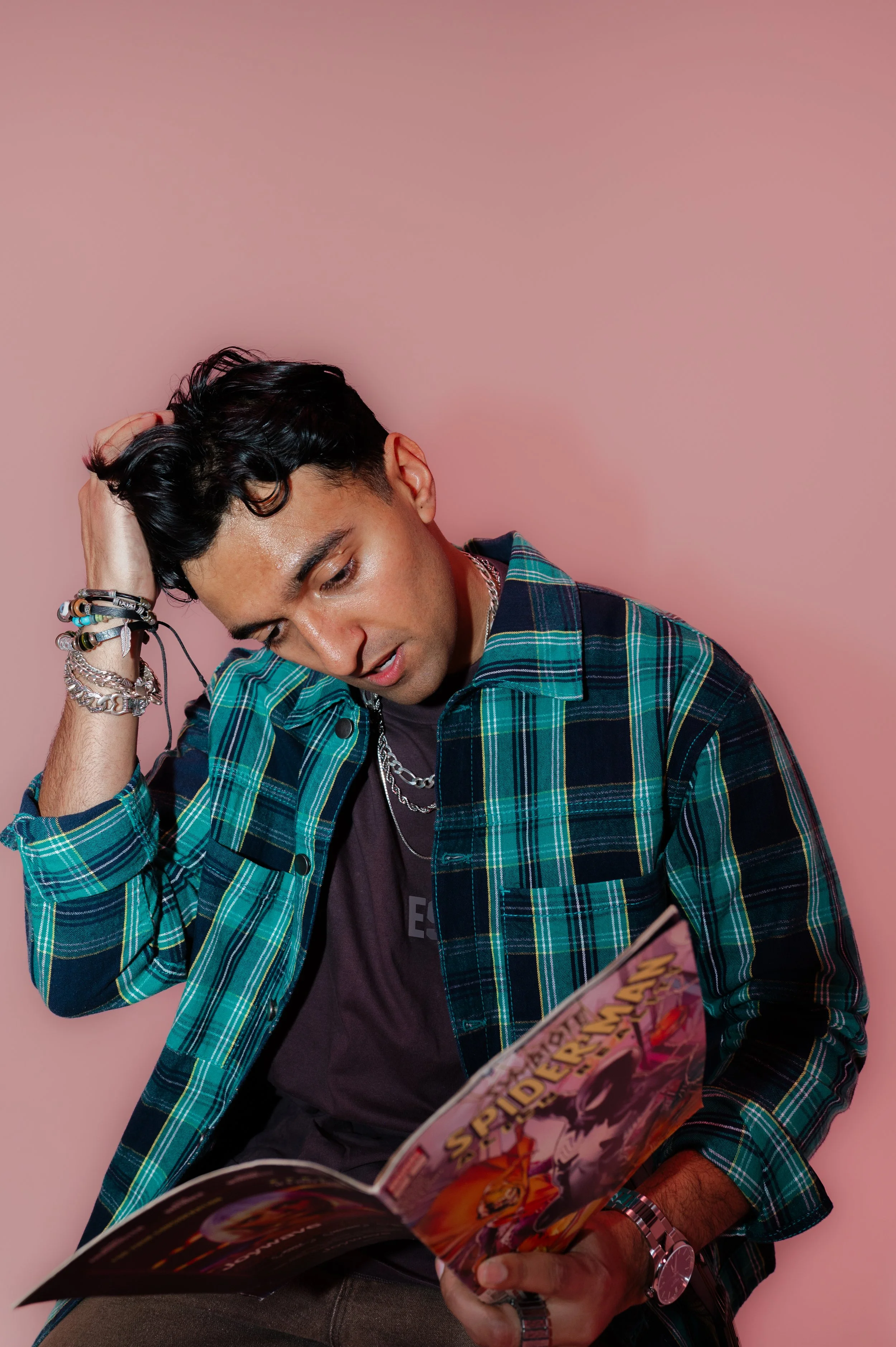 A young man with dark, wavy hair wearing a blue-green plaid shirt, multiple bracelets, and a wristwatch, is sitting against a pink background, holding and looking at a comic book titled 'SPIDER-MAN'.