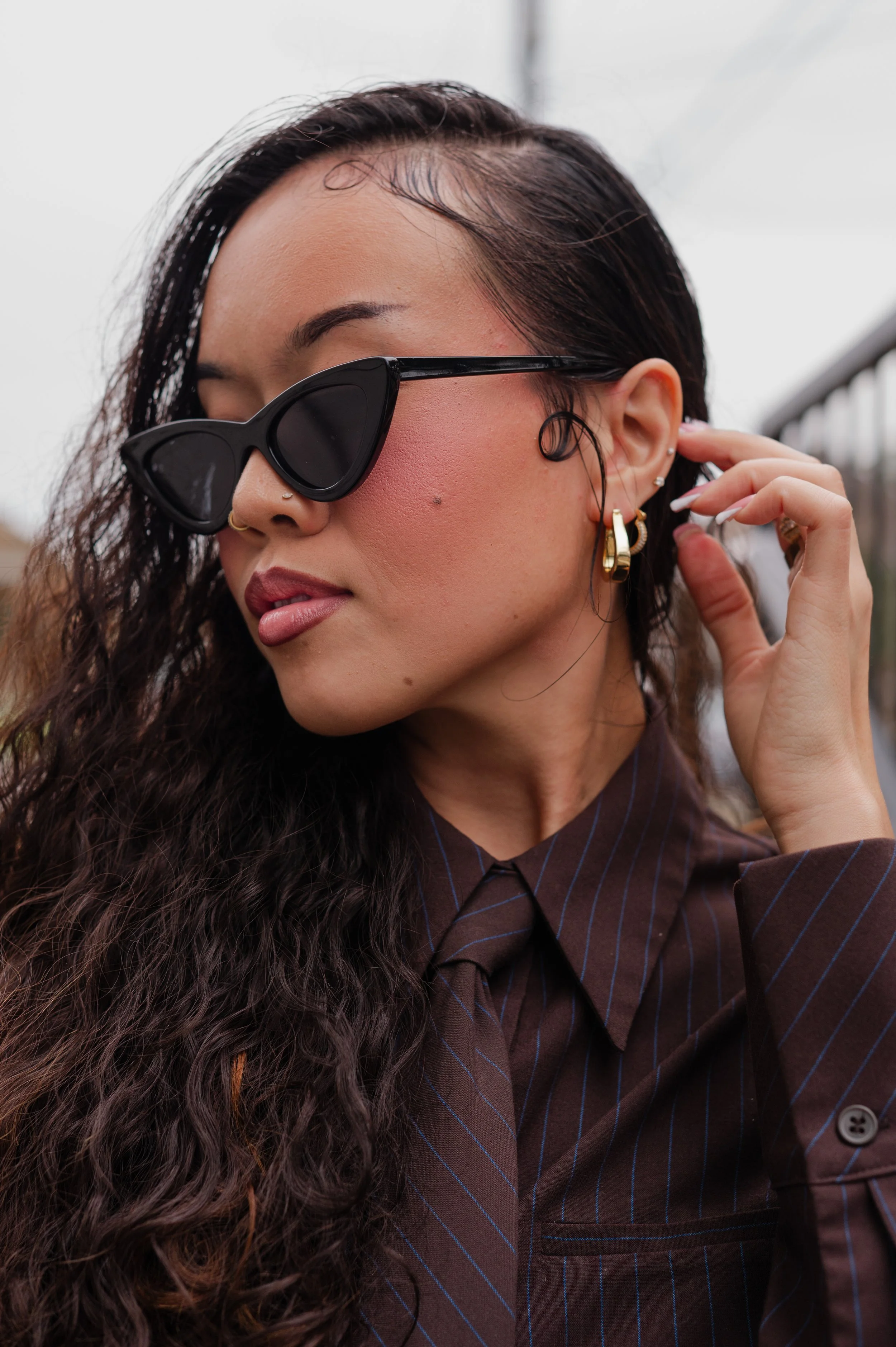 A woman with long, curly brown hair wearing black sunglasses, gold hoop earrings, and a brown pinstripe shirt, adjusting her earring outdoors with an overcast sky.