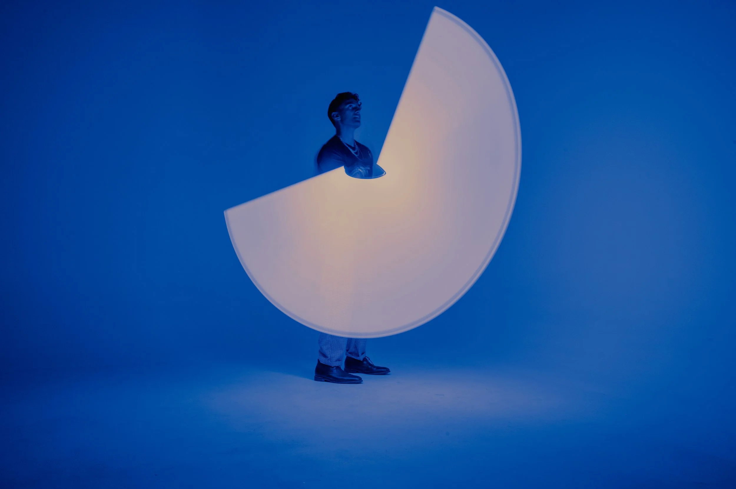 Person standing with a large, circular, reflector-like object in front of their face against a blue background.