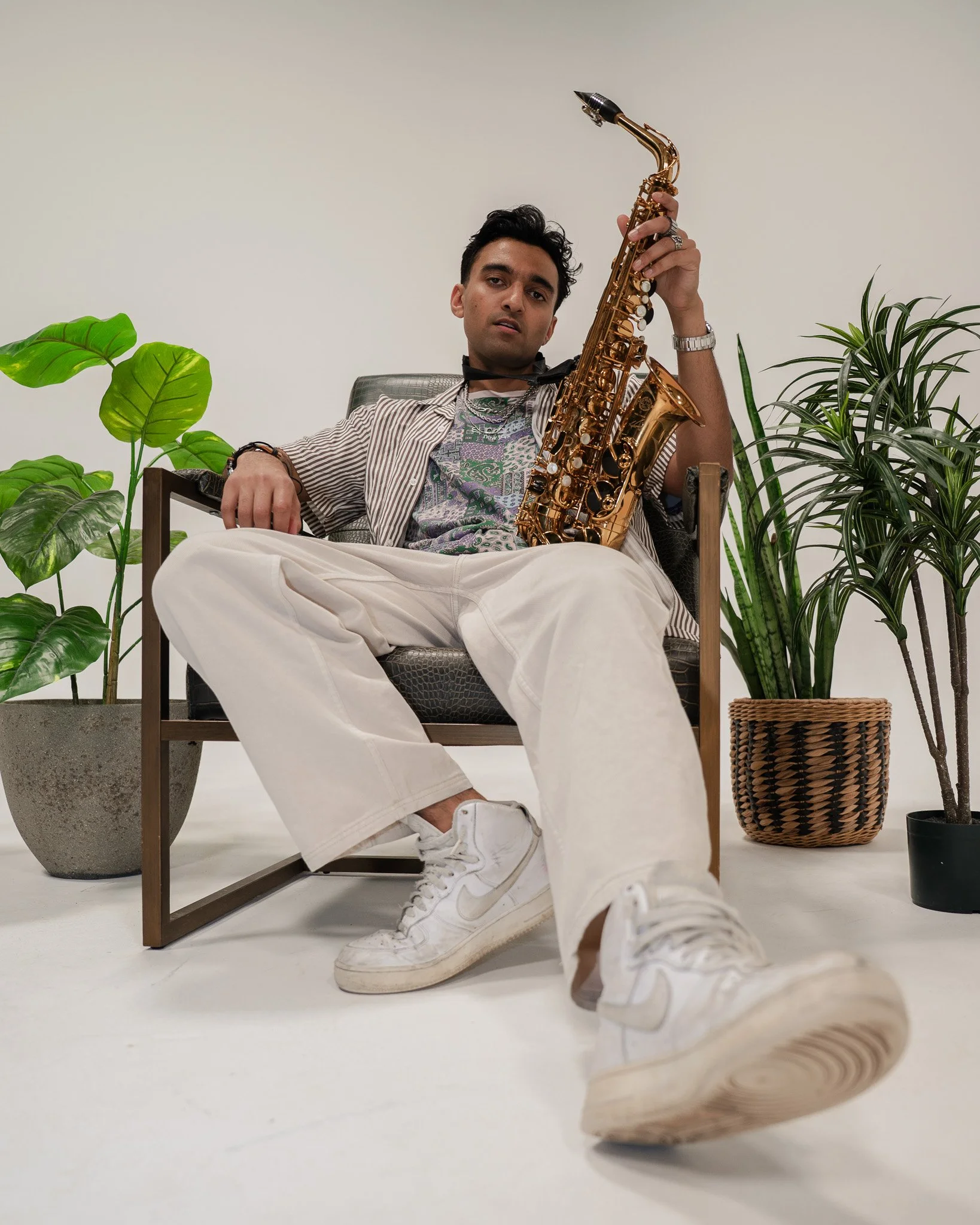 A man sitting on a chair with plants on either side, holding a gold saxophone, wearing a patterned T-shirt, striped shirt, white pants, and white sneakers.