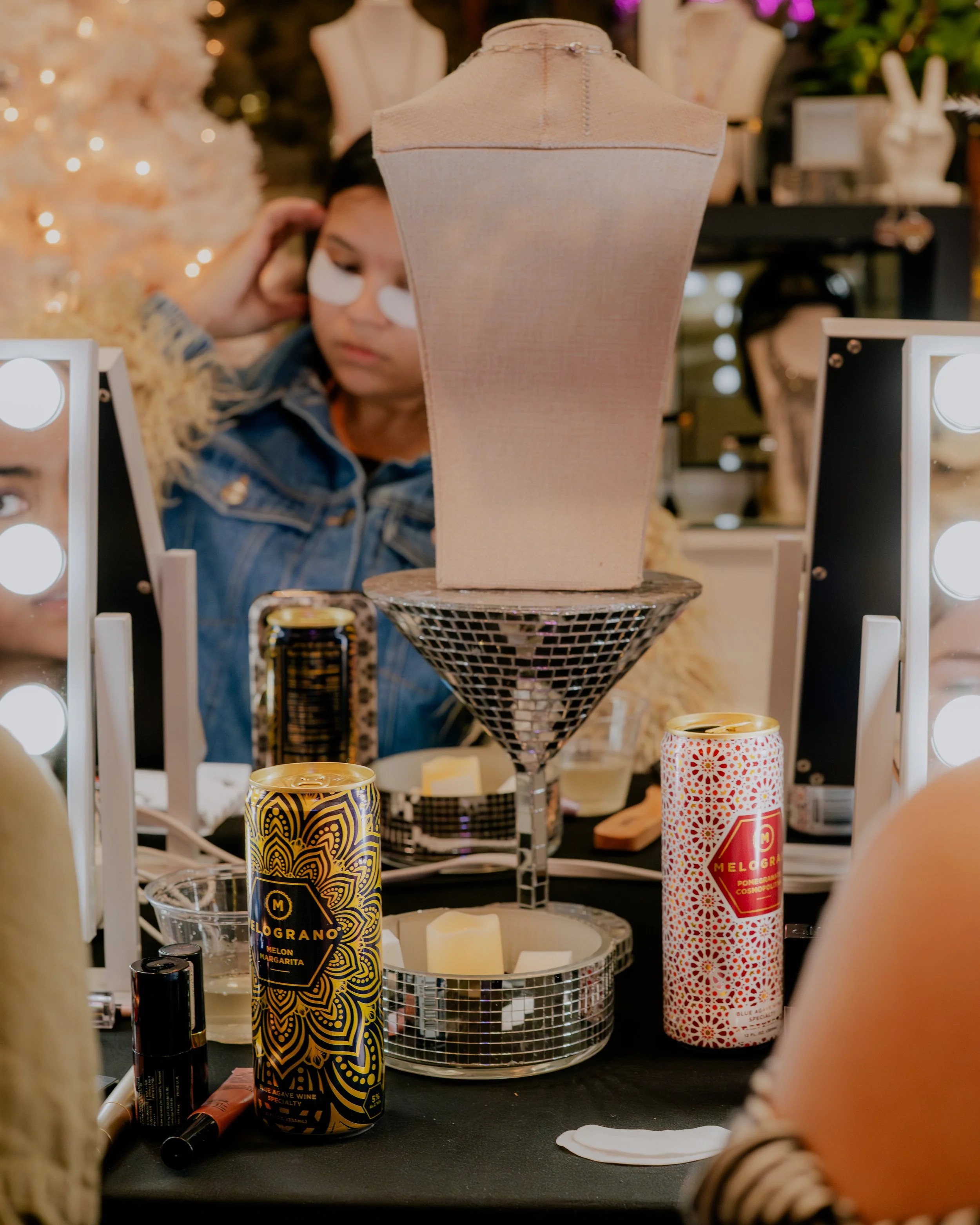 A makeup display at a store or event featuring a mannequin torso in the background, with candles, makeup products, and cans of Melograno beverage on a table in the foreground. The scene is decorated with festive lighting and Christmas decorations.