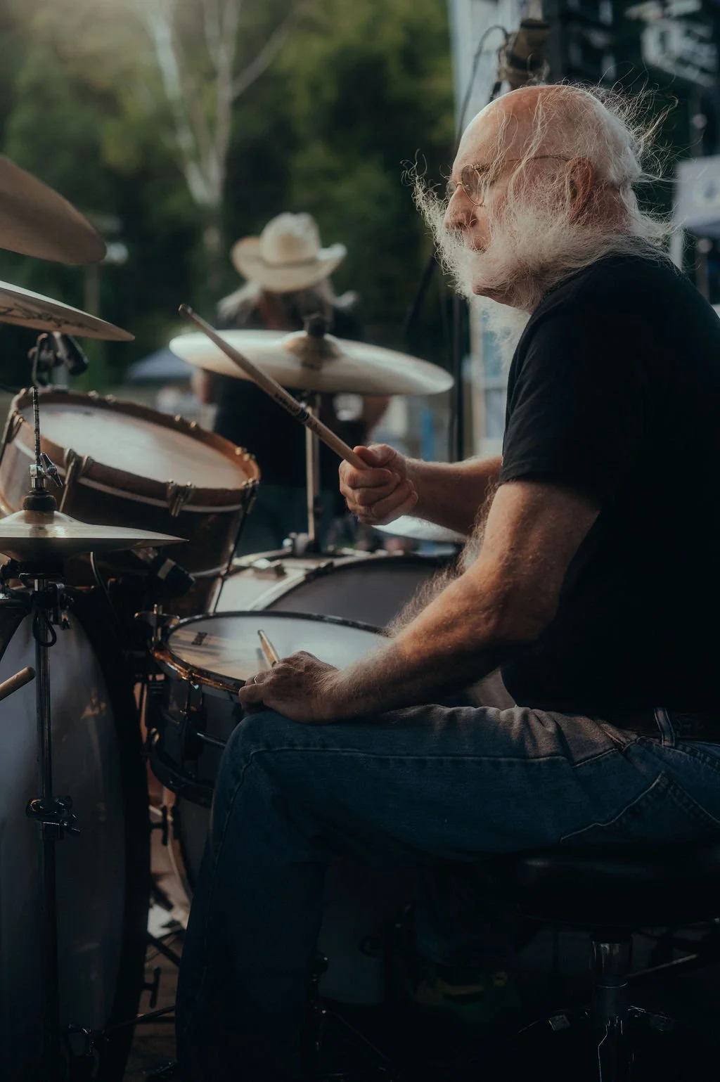 An elderly man with long white hair and beard playing drums outdoors during daytime, wearing glasses and a black shirt, with a woman in a cowboy hat in the background.