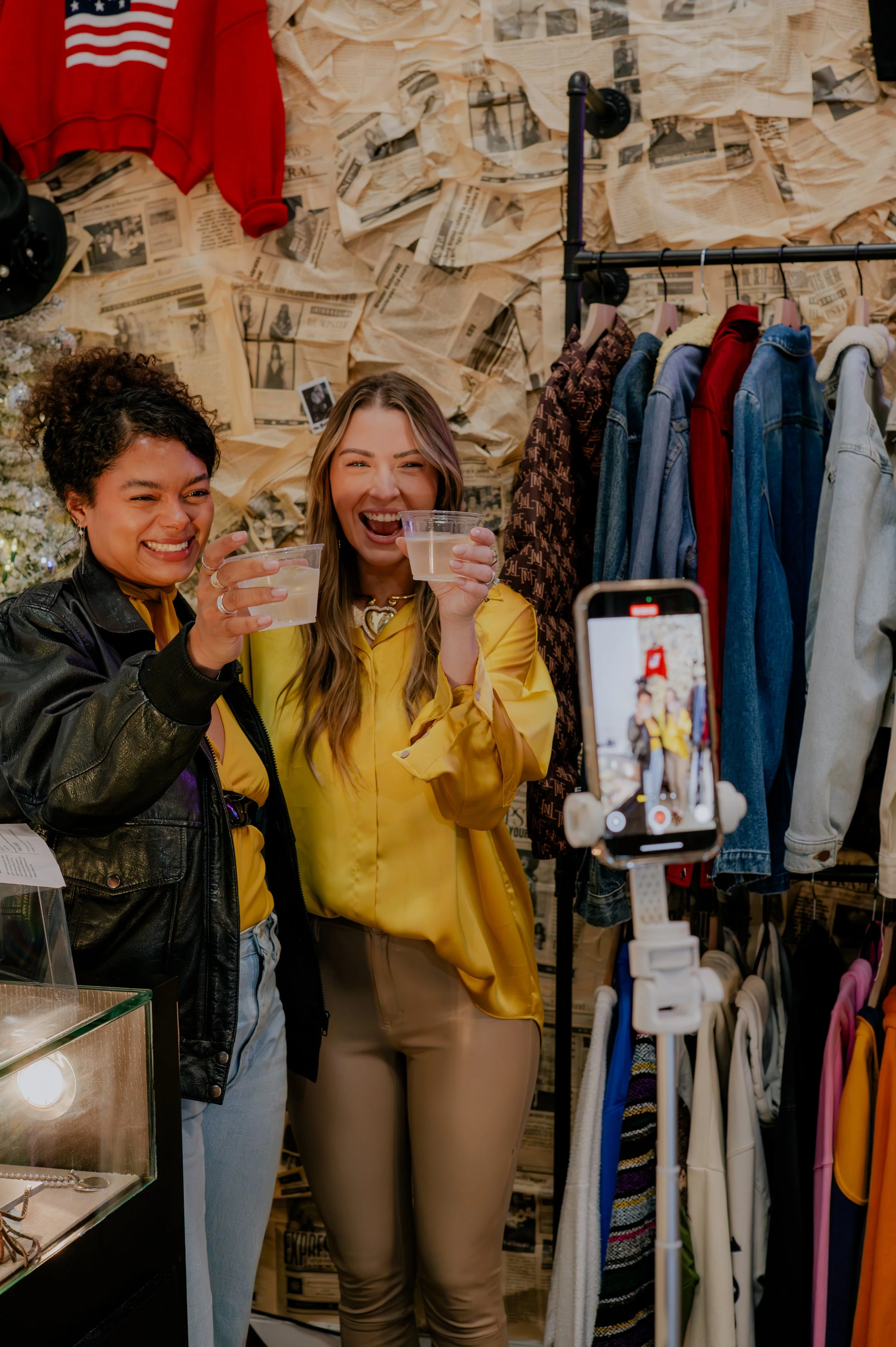 Two women smiling and clinking plastic cups with drinks in a clothing store, with clothes hanging on racks and newspaper wallpaper in the background, and a person taking their photo.