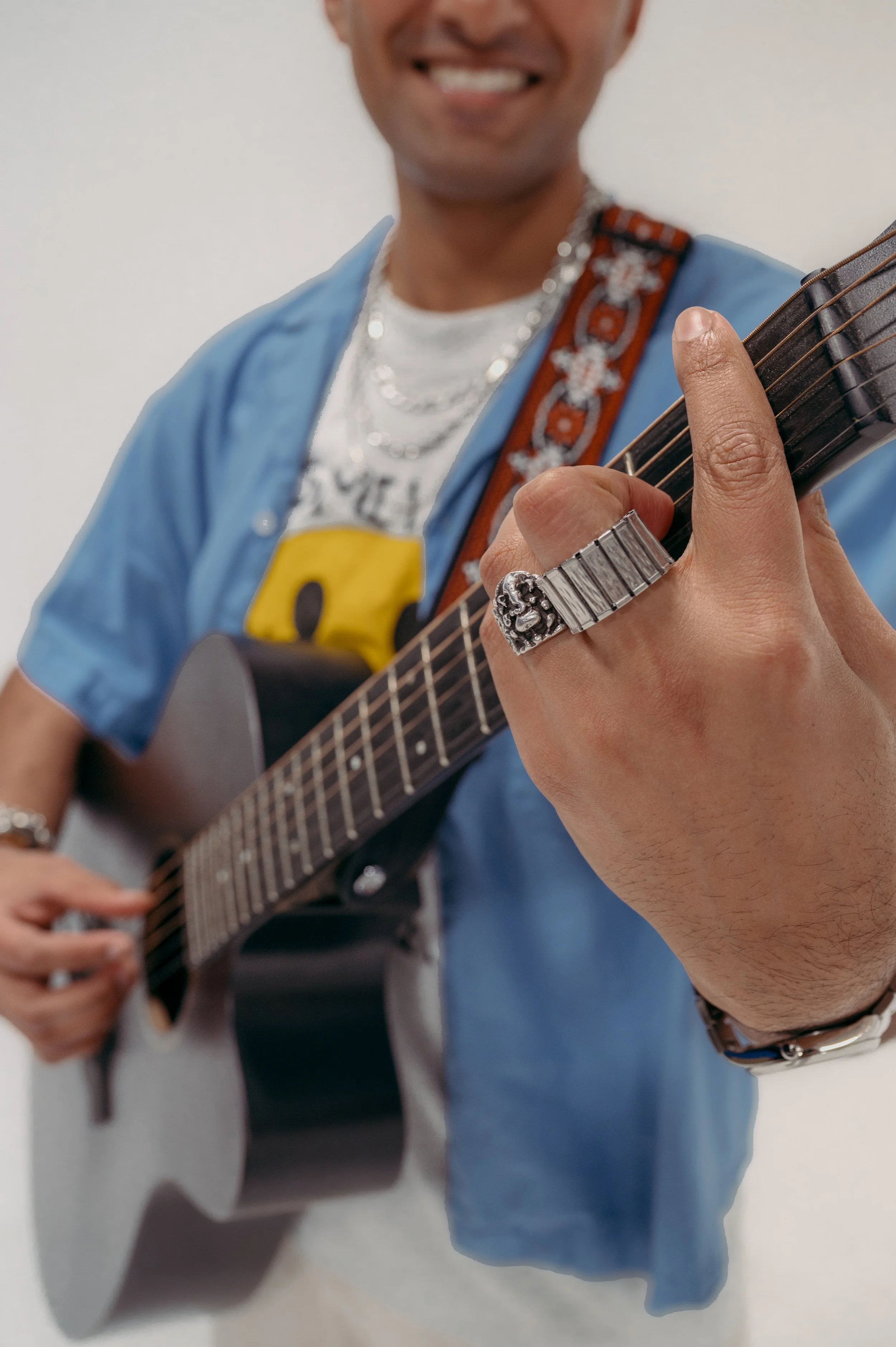 A person playing an acoustic guitar, wearing a blue jacket, a silver chain necklace, and multiple rings. The person is smiling and standing against a plain white background.