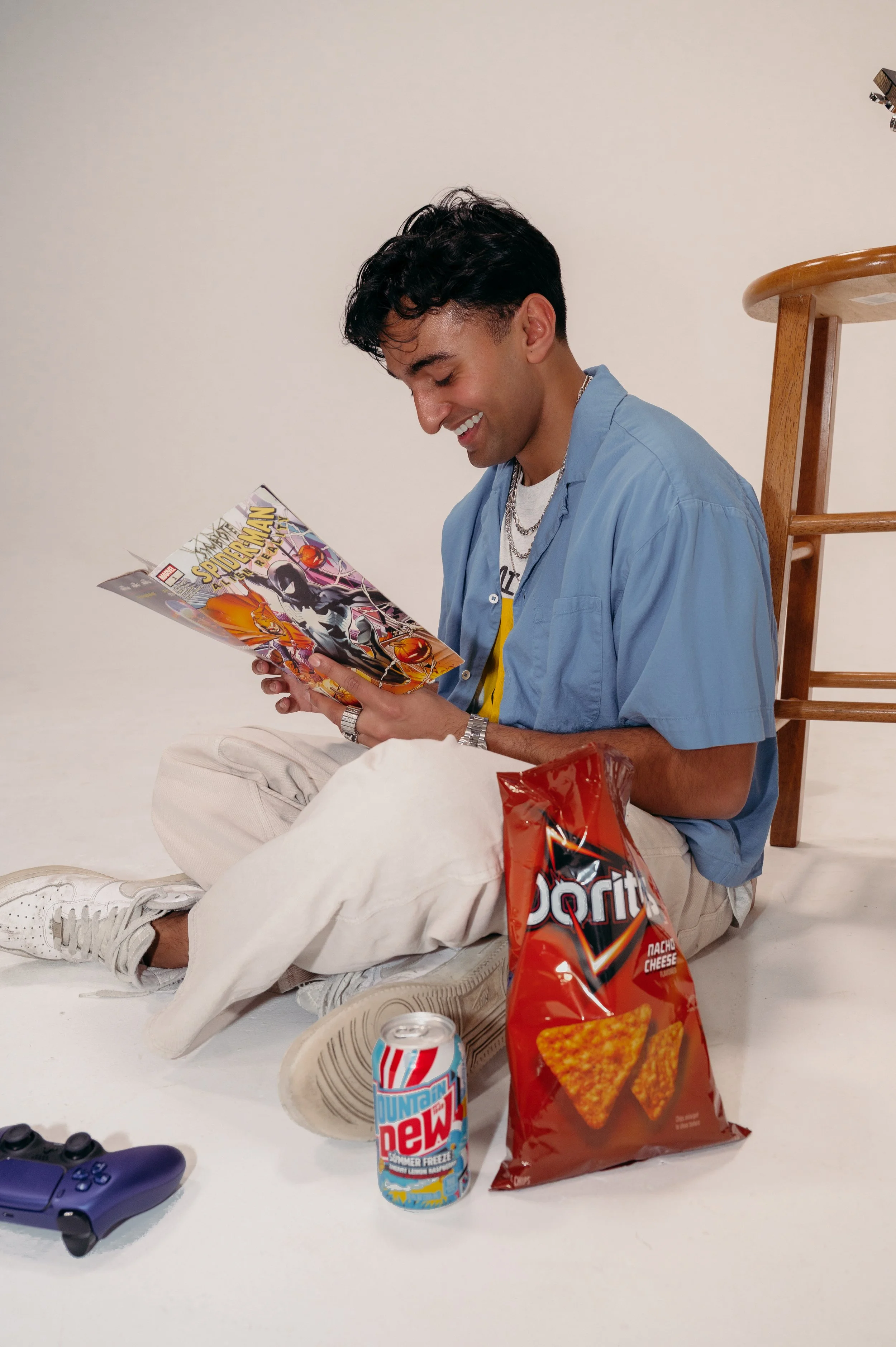 A young man sitting on the floor, smiling and reading a comic book, with snacks and drinks around him, including a bag of Doritos Nacho Cheese chips and a Mountain Dew soda.