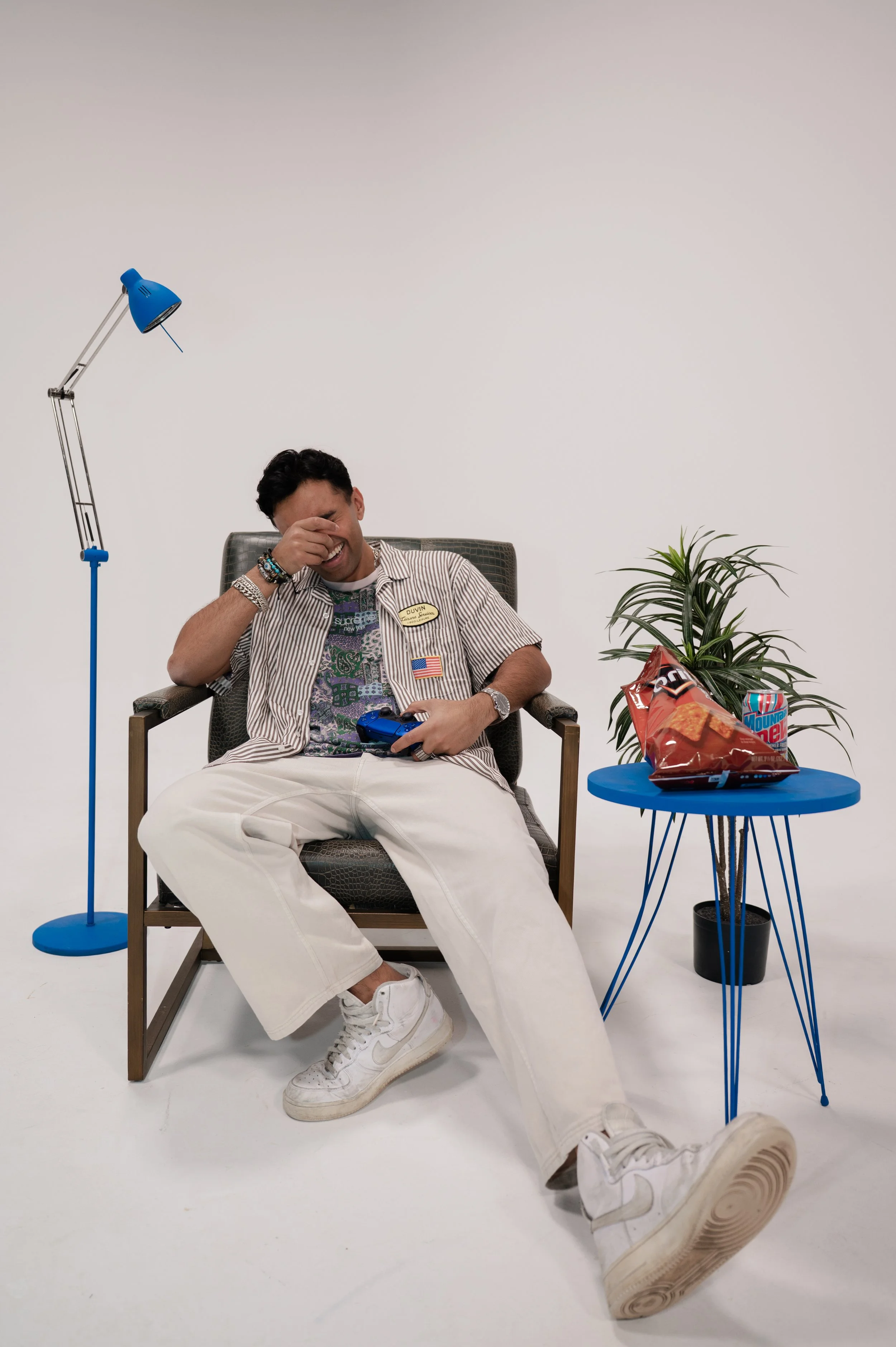 A young man with dark hair is sitting in a chair, laughing and covering his face with one hand. He is dressed in a striped shirt, baggy white pants, and white sneakers. On his chest, he has multiple colorful badges and an American flag patch. Next to