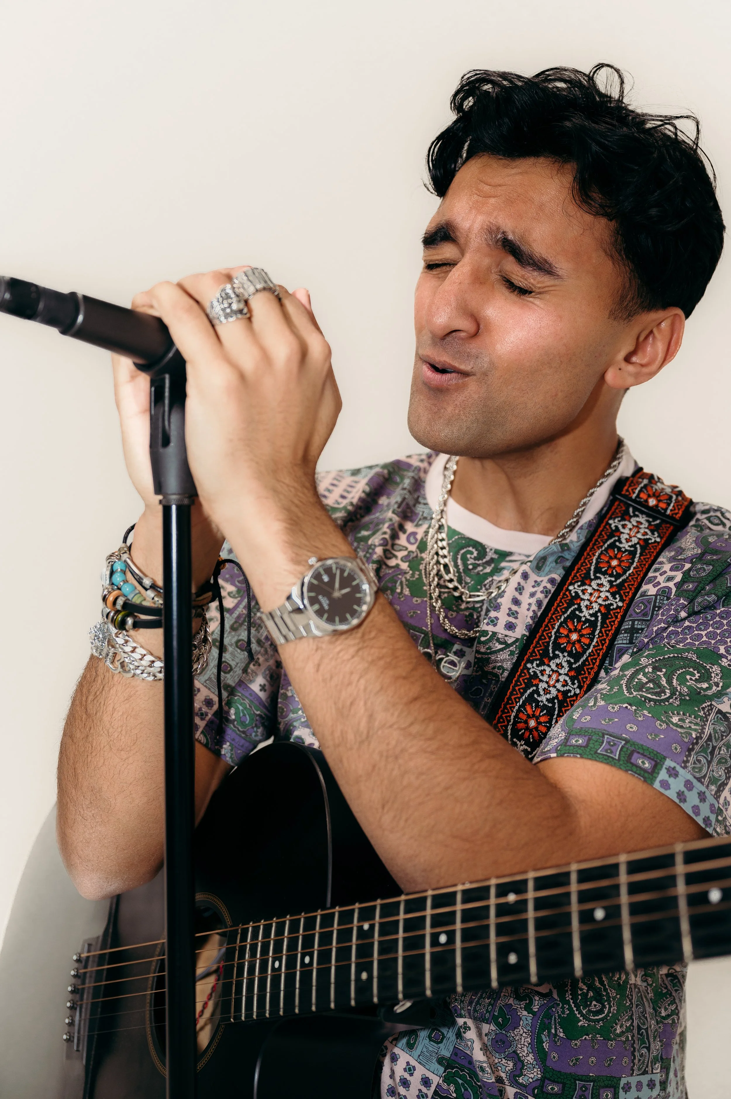 A man with dark, curly hair singing into a microphone while playing an acoustic guitar. He is wearing multiple bracelets, a watch, and layered necklaces, with a patterned shirt.