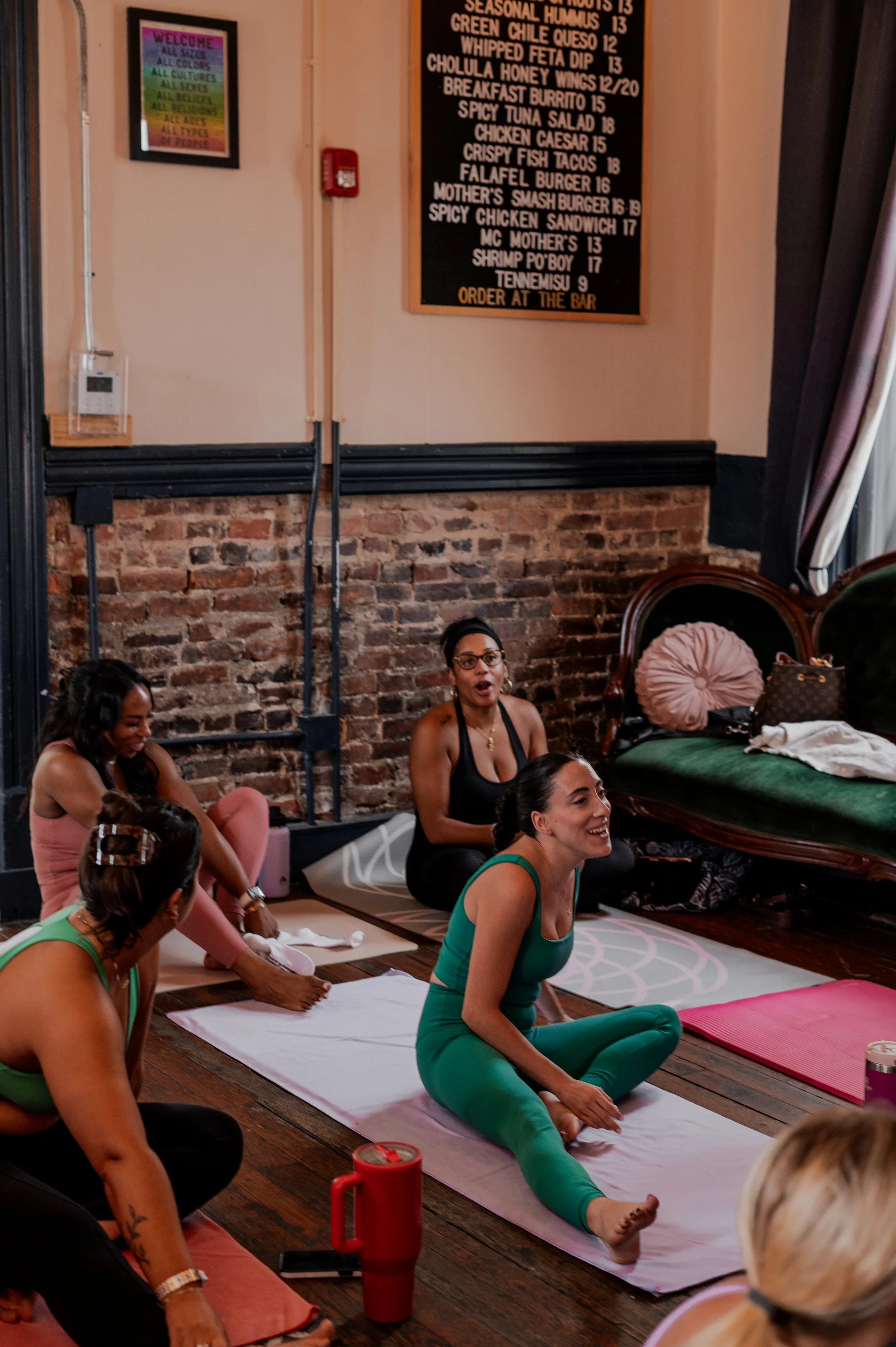 Women participating in a yoga class, sitting on yoga mats in a studio with exposed brick wall and a blackboard menu visible in the background.
