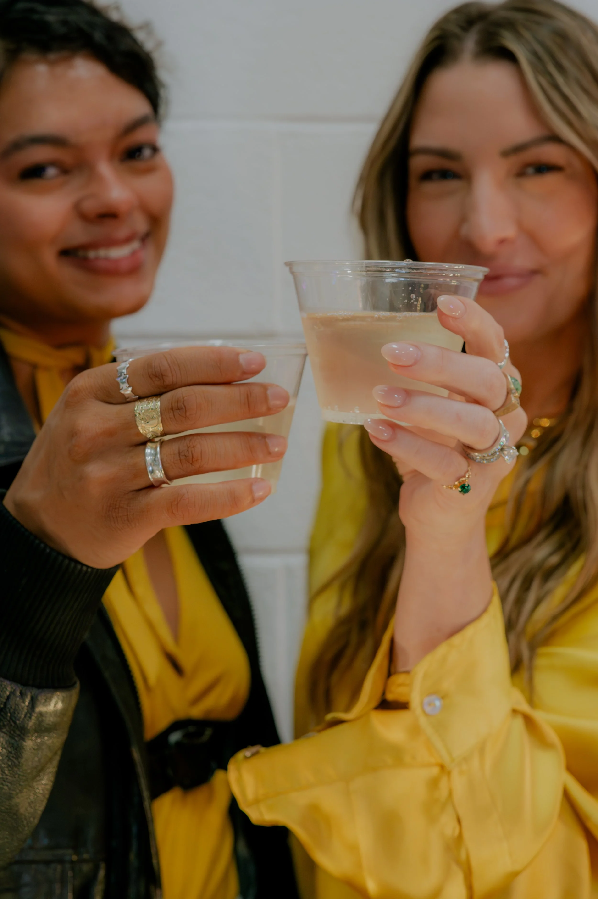 Two women holding glasses of beverage, smiling and celebrating together, standing against a plain wall.