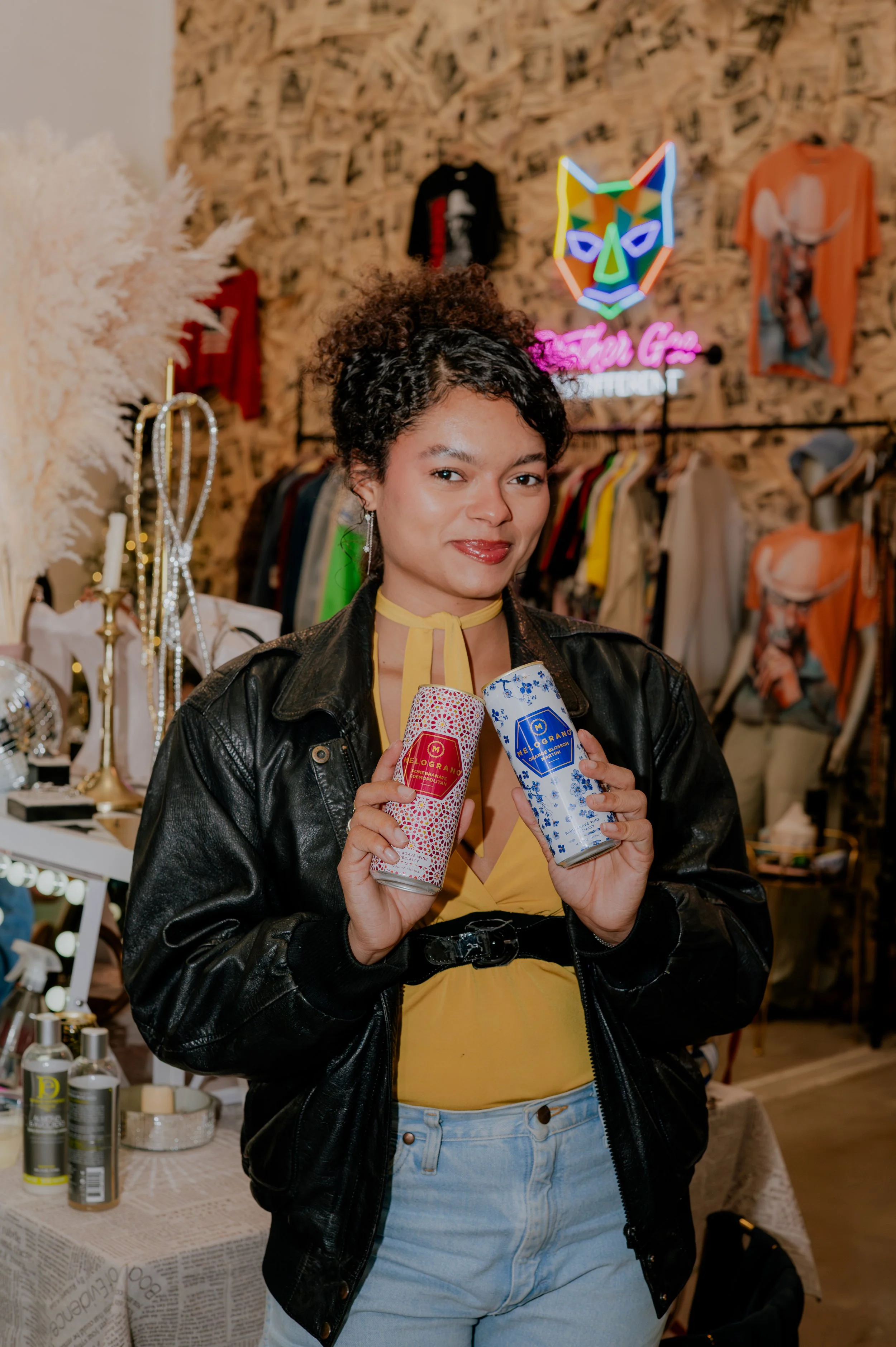 A woman with curly hair, wearing a yellow top and a black leather jacket, holding two cans of beverage in front of a retail clothing store with clothing racks and decorative items in the background.