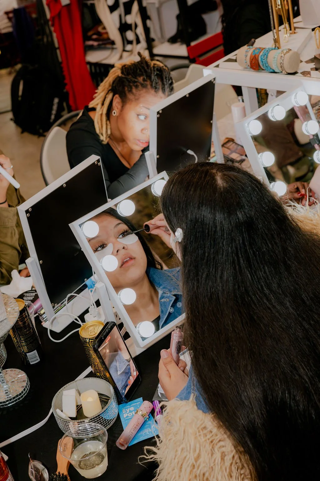 A woman is applying makeup to a woman's face using a mirror with bright lights around the edges, in a busy makeup studio.