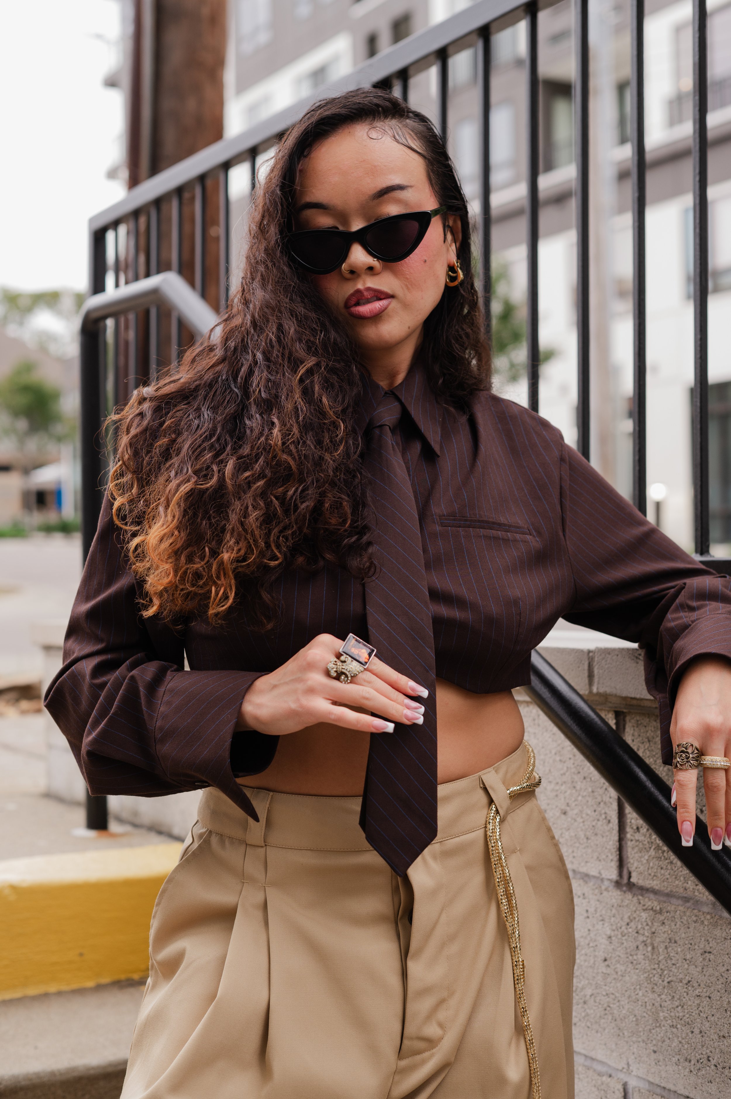 A woman with long curly hair wearing sunglasses, a dark brown open shirt, and beige high-waisted pants, standing outdoors near a metal railing and a brick wall building in the background.