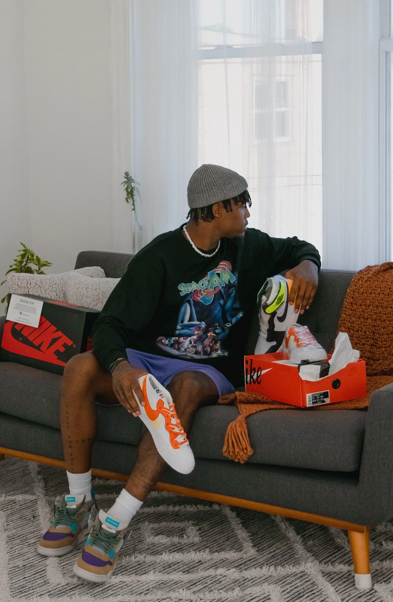 A young man sitting on a gray couch, holding a sneaker in each hand, with shoe boxes and wrapping paper around him, in a room with sheer curtains and a carpeted floor.