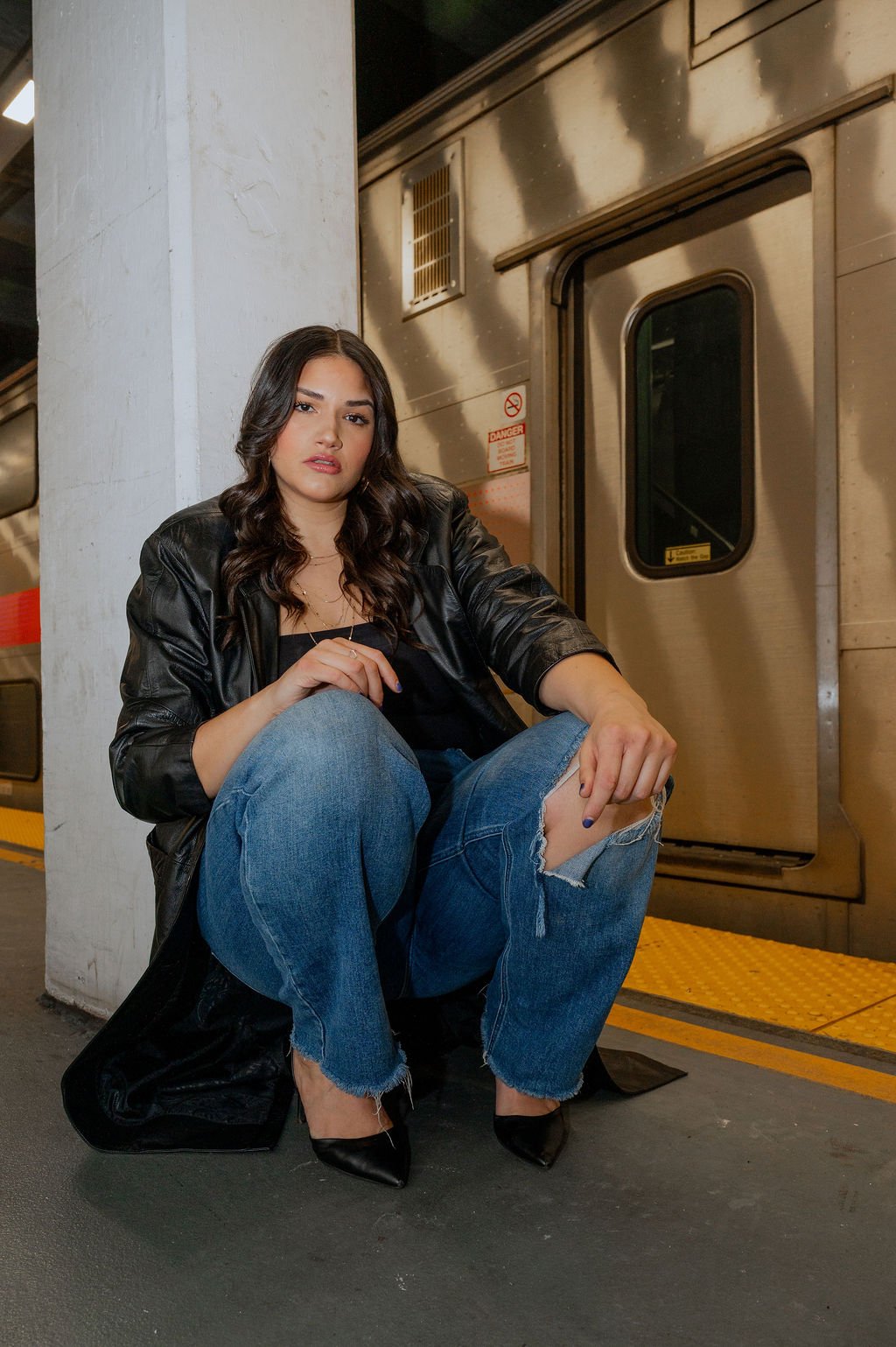 A woman with dark wavy hair sitting on the ground in a subway station, wearing a black leather jacket, ripped jeans, and black pointed shoes, next to a metallic subway door.