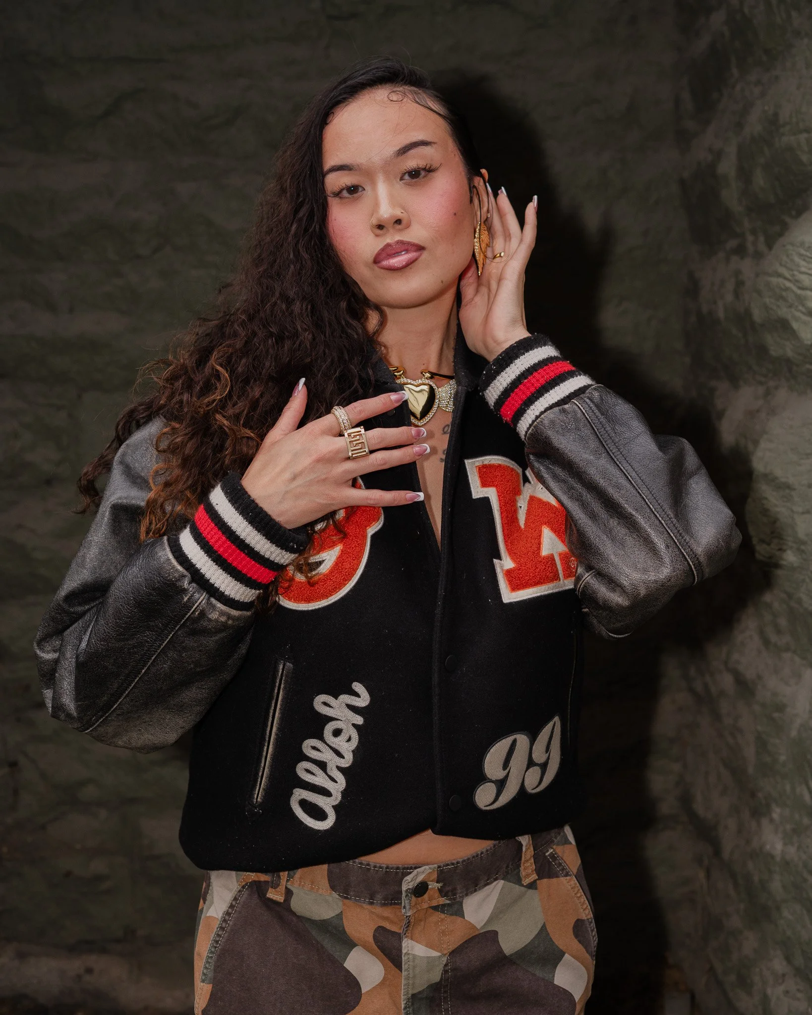 A woman with long curly hair, gold earrings, and layered necklaces posing with her hand near her face against a dark stone wall. She is wearing a black and gray varsity jacket with red and white accents over camouflage pants.