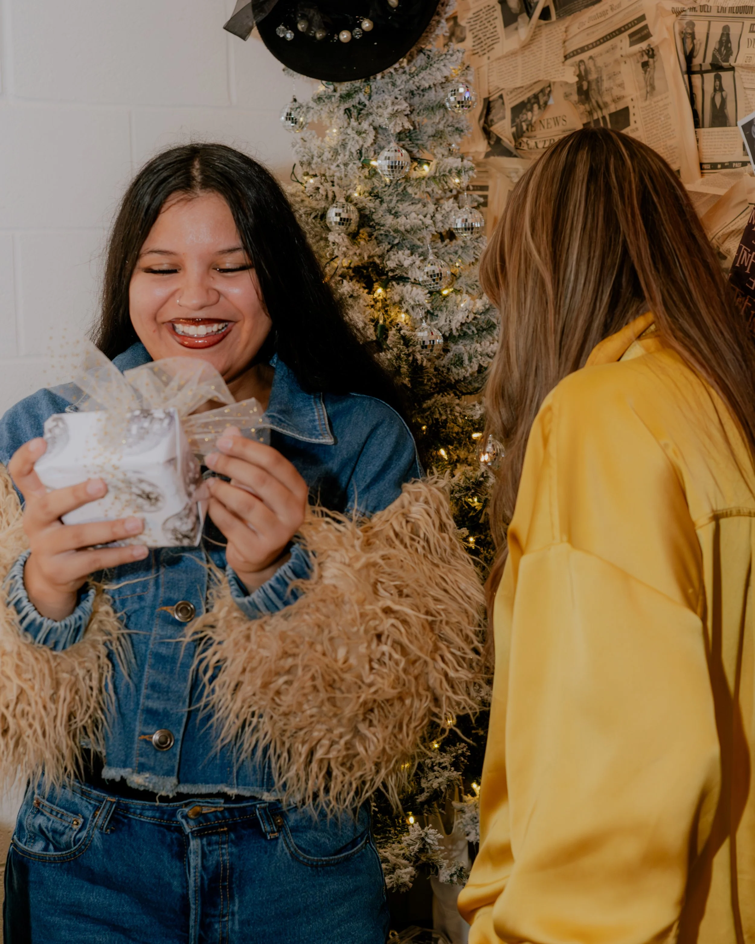 Two women exchanging a gift in front of a decorated Christmas tree with white snow-like branches and shiny silver ornaments. One woman has dark hair, is smiling, and is wearing a denim jacket over a furry coat. The other woman has light brown hair an