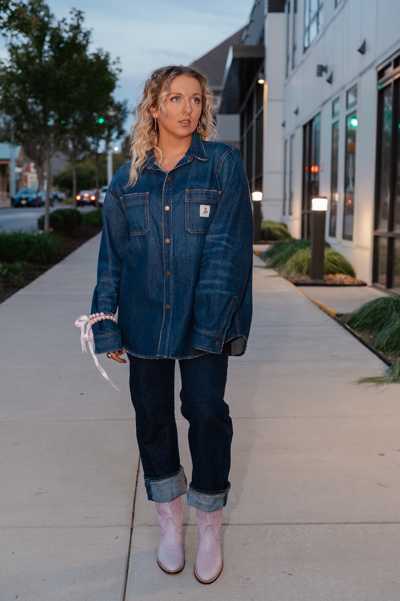 A woman with blonde, curly hair, wearing a denim jacket, dark jeans, and pink cowboy boots, walking on a sidewalk during dusk.