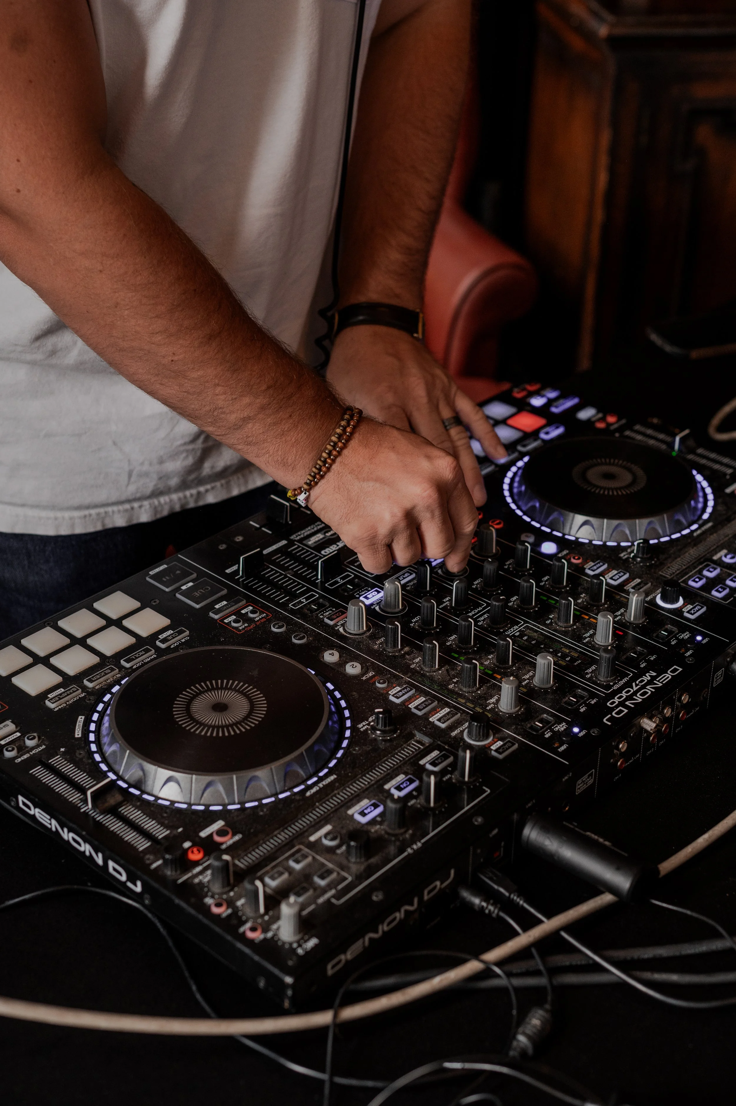 A DJ wearing a white shirt and bracelets, operating a Denon DJ controller in a dimly lit setting.