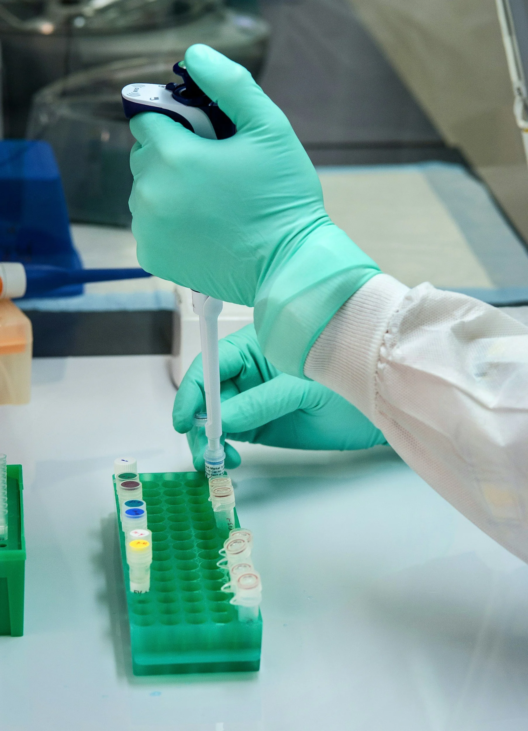 A scientist wearing green gloves and a white lab coat using a pipette to transfer liquid into small test tubes in a laboratory setting.
