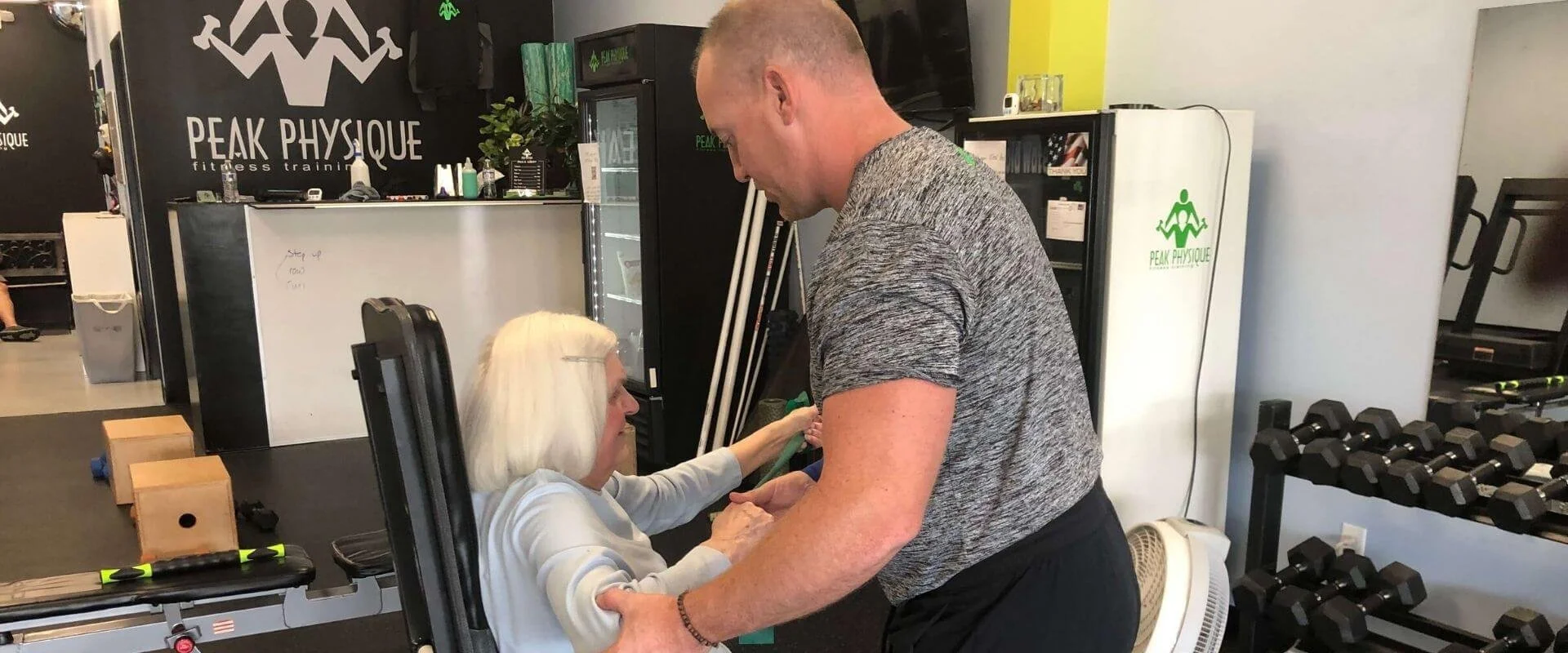 An elderly woman with white hair sitting on fitness equipment, talking to a man standing in front of her inside a gym named Peak Physique. The gym has black walls, fitness equipment, dumbbells, and a beverage refrigerator.