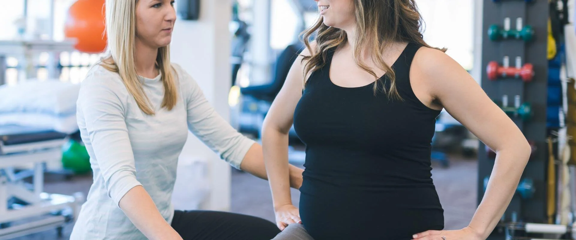 A physical therapist or trainer assists a woman in a black tank top with an exercise or adjustment in a gym or rehabilitation center.