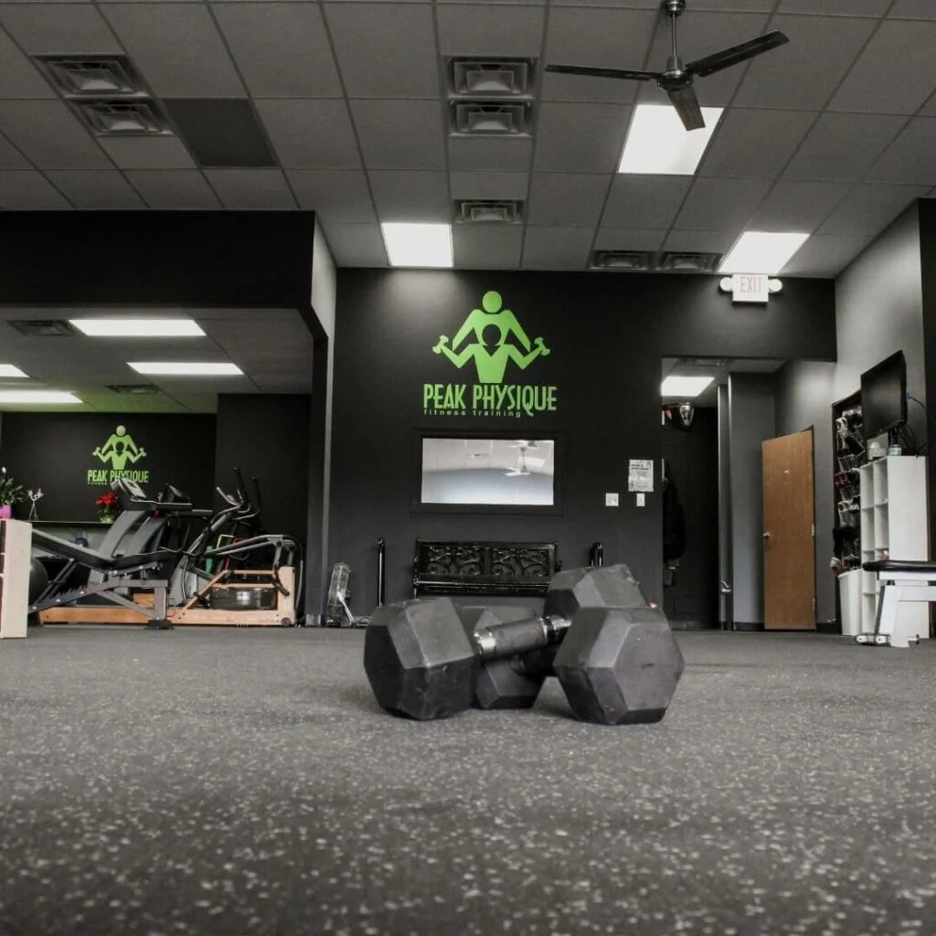 Fitness gym with a pair of black dumbbells on a speckled gray floor, PEAK PHYSIQUE fitness training sign on the wall, and gym equipment in the background.