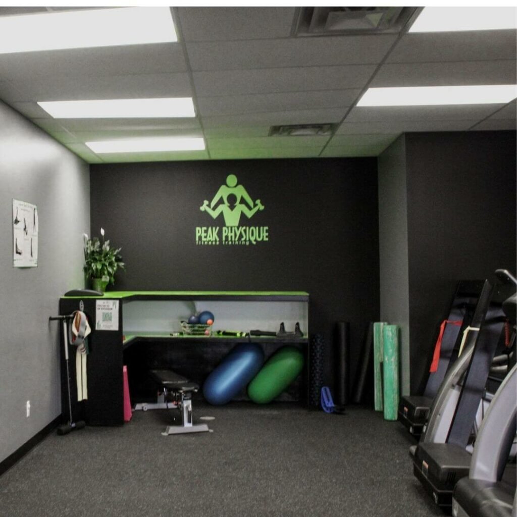 An interior view of a fitness gym with PEAK PHYSIQUE lighting on the wall, workout equipment including foam rollers, exercise mats, and workout machines visible.