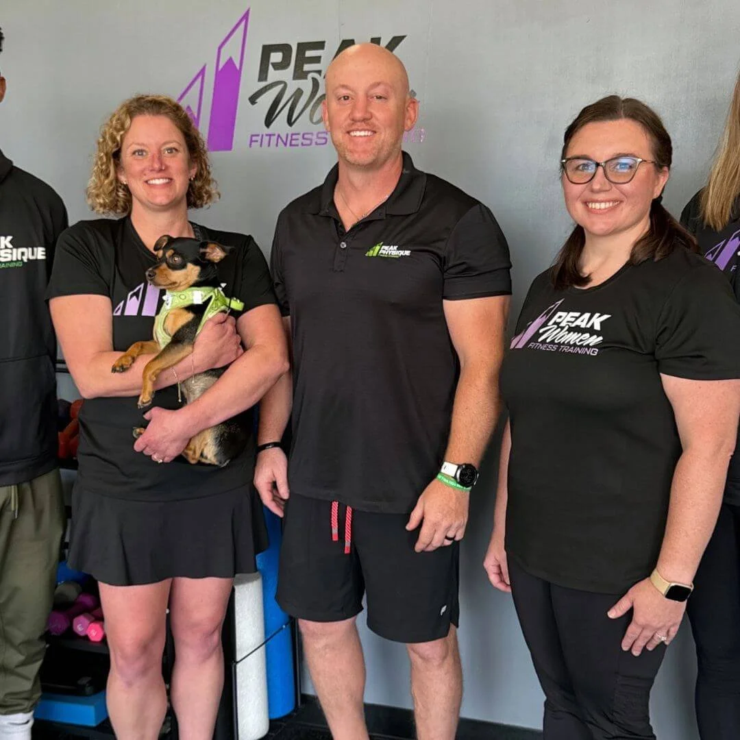 Group of four people standing together in a fitness gym, smiling at the camera. One woman is holding a small dog. All are wearing black workout shirts with the Peak Women Fitness Training logo.