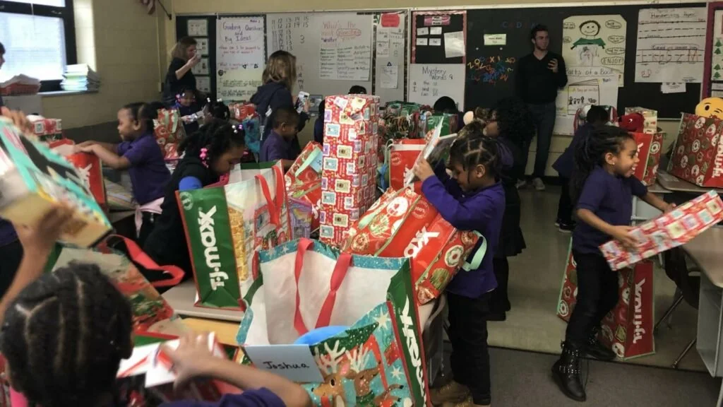 A classroom filled with young children opening Christmas presents, with teachers or adults assisting. Many large, festive gift bags and boxes are spread across tables and desks. Children are wearing purple uniforms and appear excited during a holiday gift exchange.
