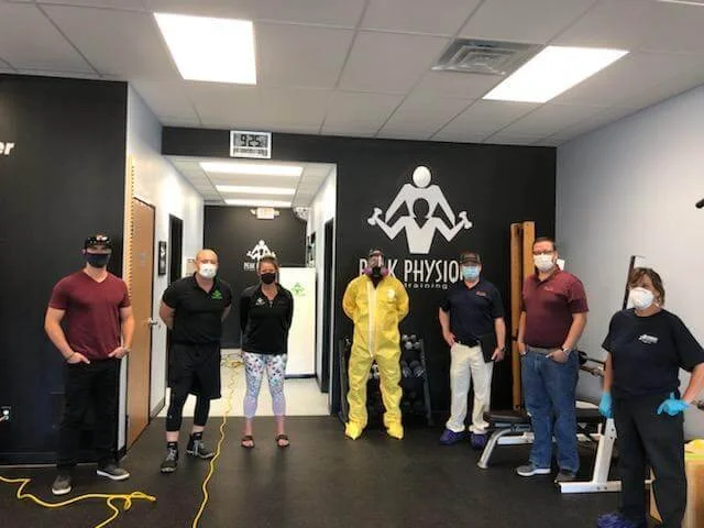 Seven people standing in a physical therapy clinic with black walls, showing staff in casual and medical attire, some wearing masks and gloves, with equipment and a logo on the wall that says 'Black Physio.'