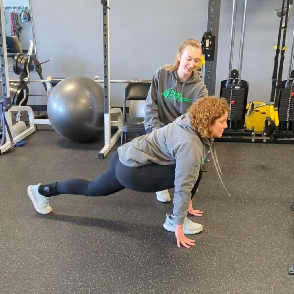A woman with curly hair in black leggings and a grey hoodie is performing a plank exercise with one leg raised, supported on her hands and toes on a gym floor. A trainer with long reddish hair in a grey hoodie watches her and provides support at a gym with various exercise equipment in the background.