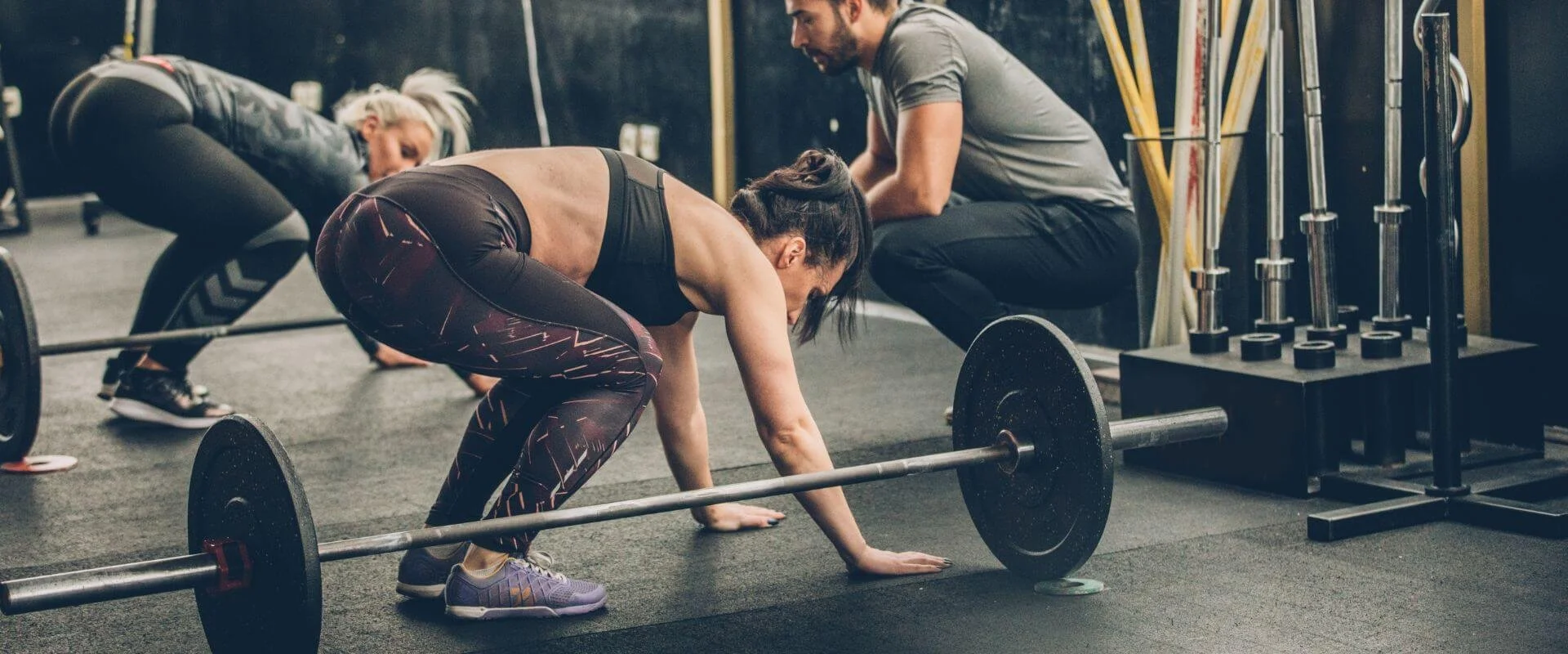Three women are performing deadlifts in a gym with a coach or trainer supervising. They are in a bent-over position, lifting barbells with weights on the floor.
