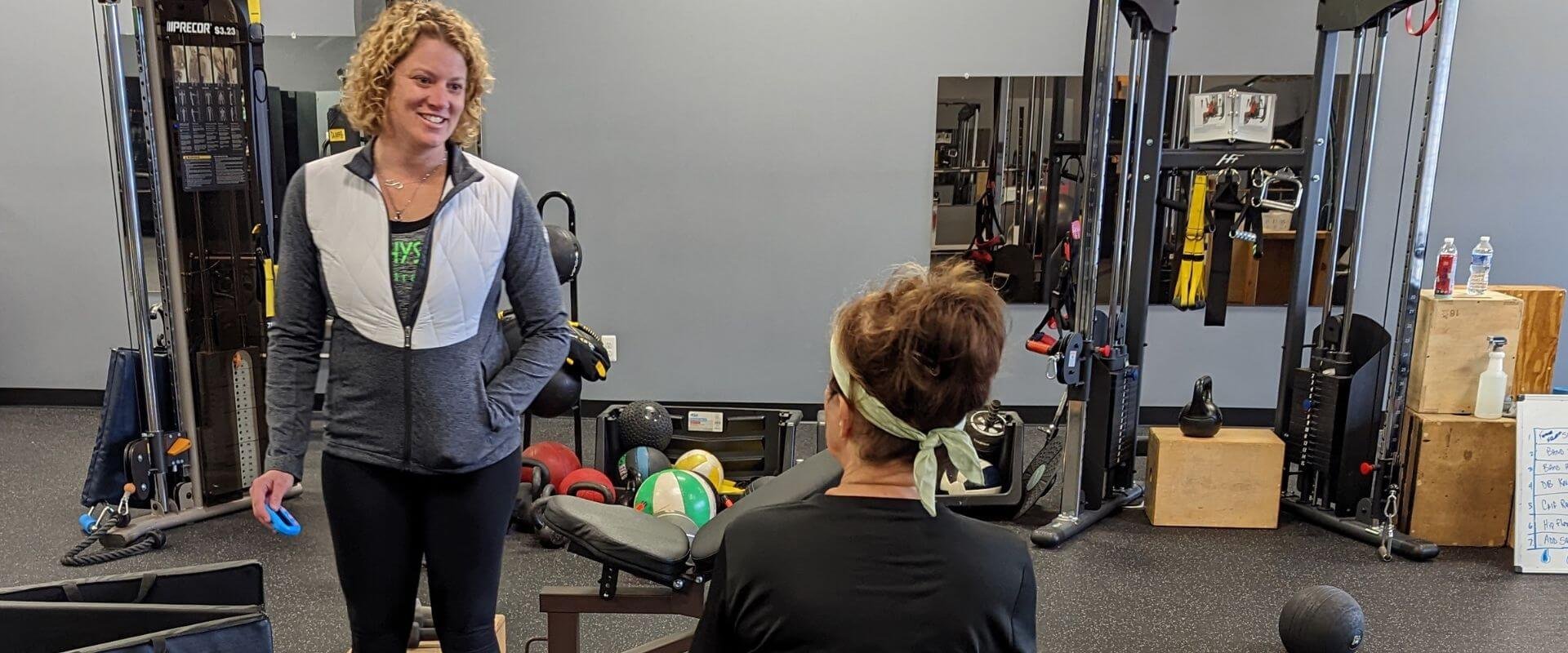 Two women in a gym. One woman is standing, smiling, holding a small exercise resistance band. The other woman is sitting and wearing workout clothes and a headband, facing the standing woman. The gym has various exercise equipment, kettlebells, medicine balls, and water bottles visible.