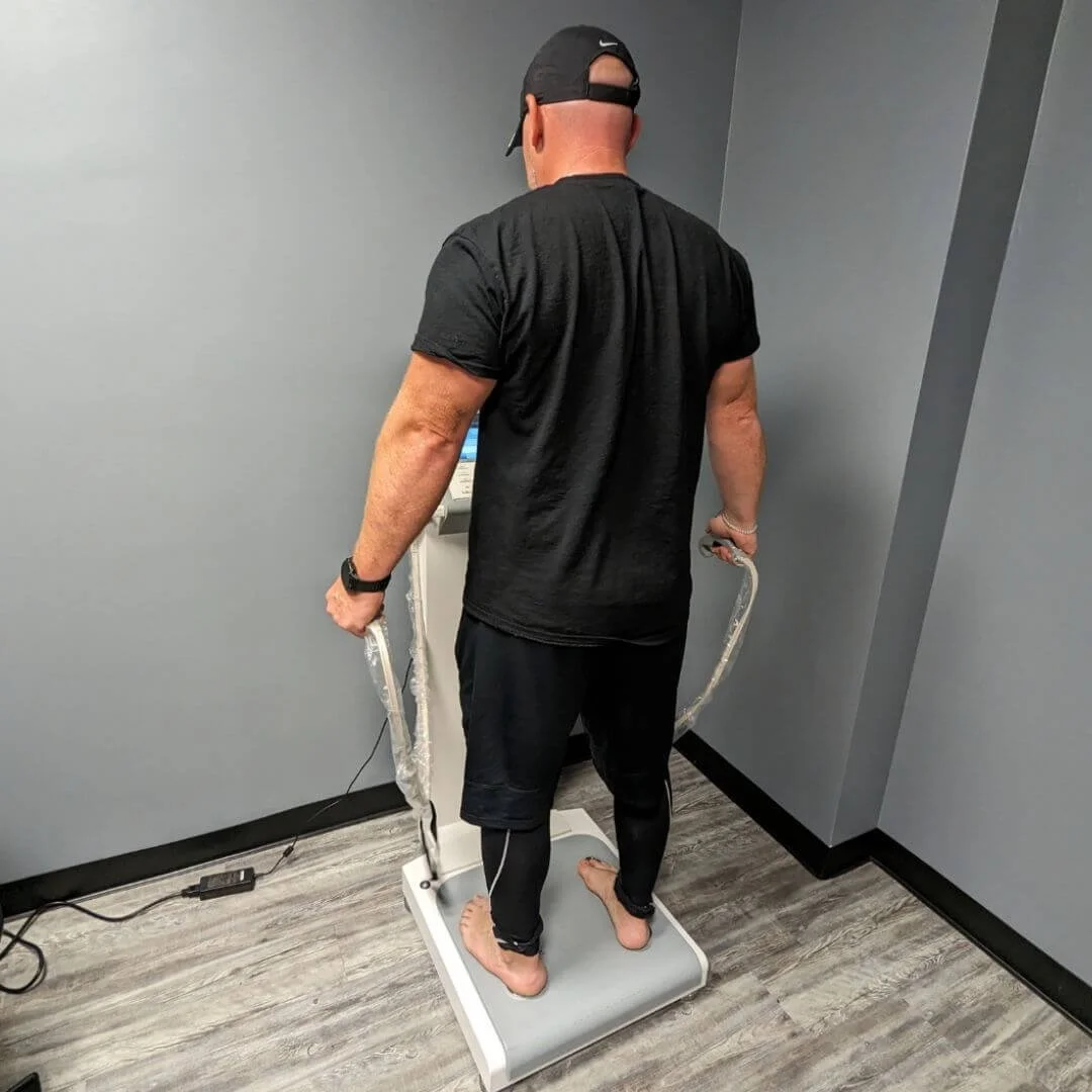A man standing barefoot on a body composition analyzer scale in a room with gray walls and hardwood floor, wearing a black t-shirt, shorts, and a black cap, holding metal hand grips connected to the device.