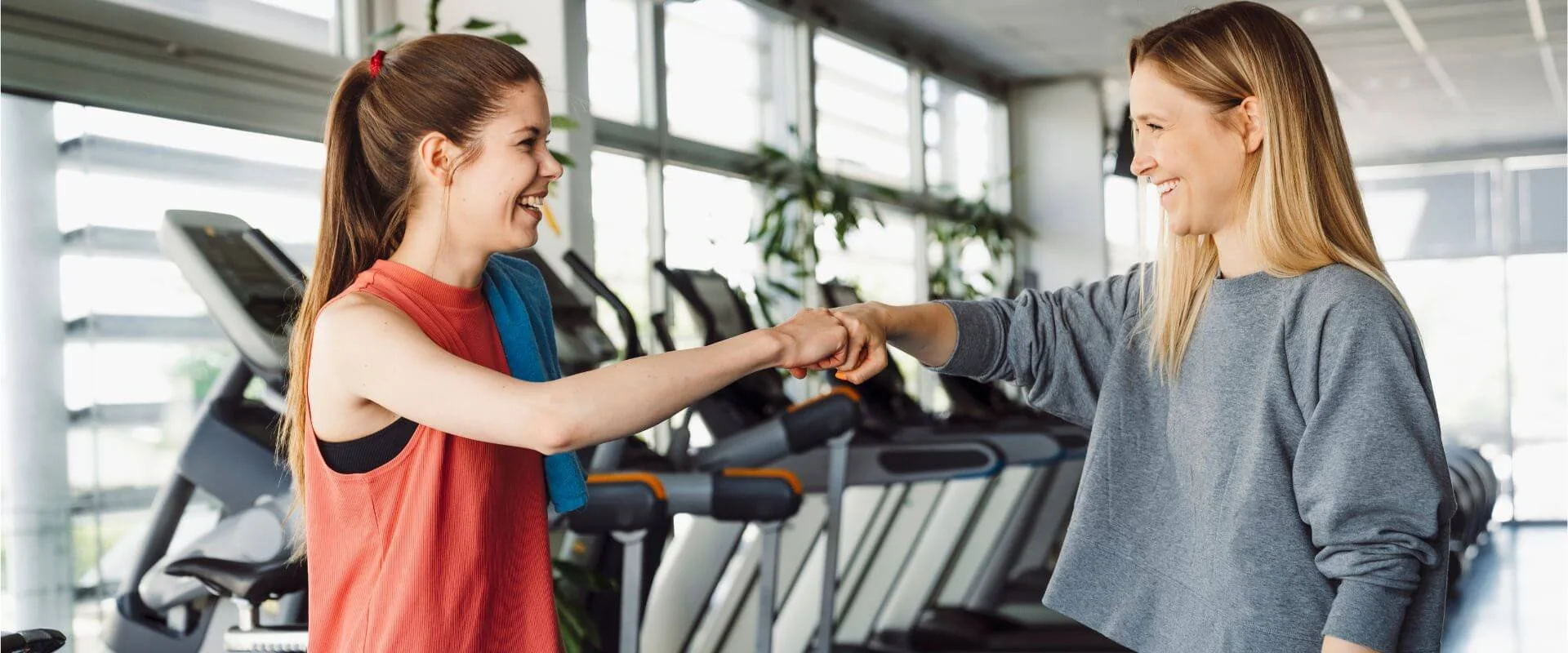 Two women smiling and fist-bumping in a gym with treadmills in the background.