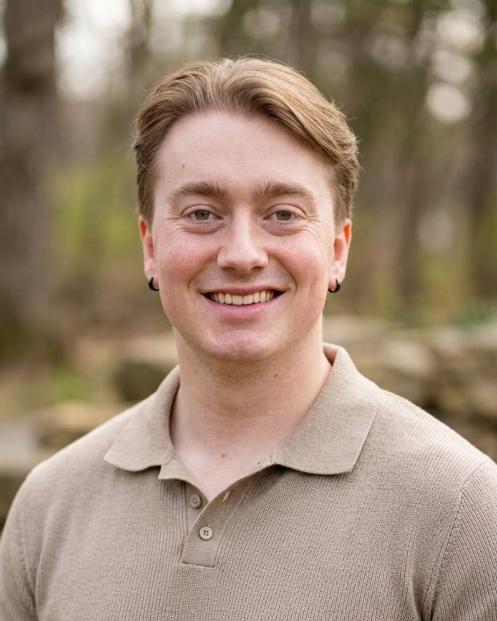 Close-up of a smiling person with short hair in an outdoor setting, wearing a beige polo shirt.