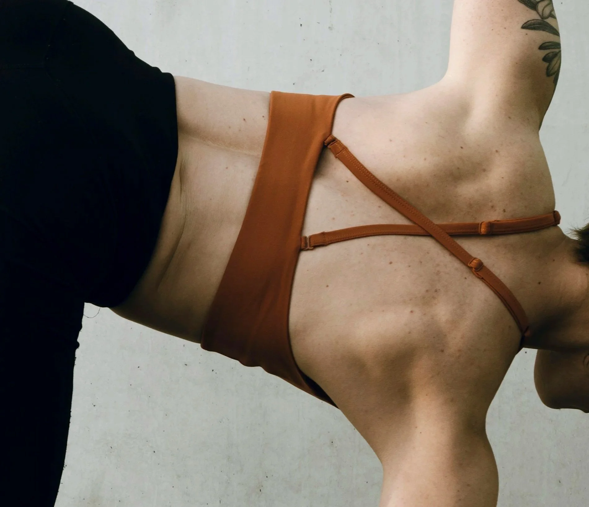 Close-up of a woman wearing black shorts and a brown bra, with a tattoo on her upper arm, against a plain white background.