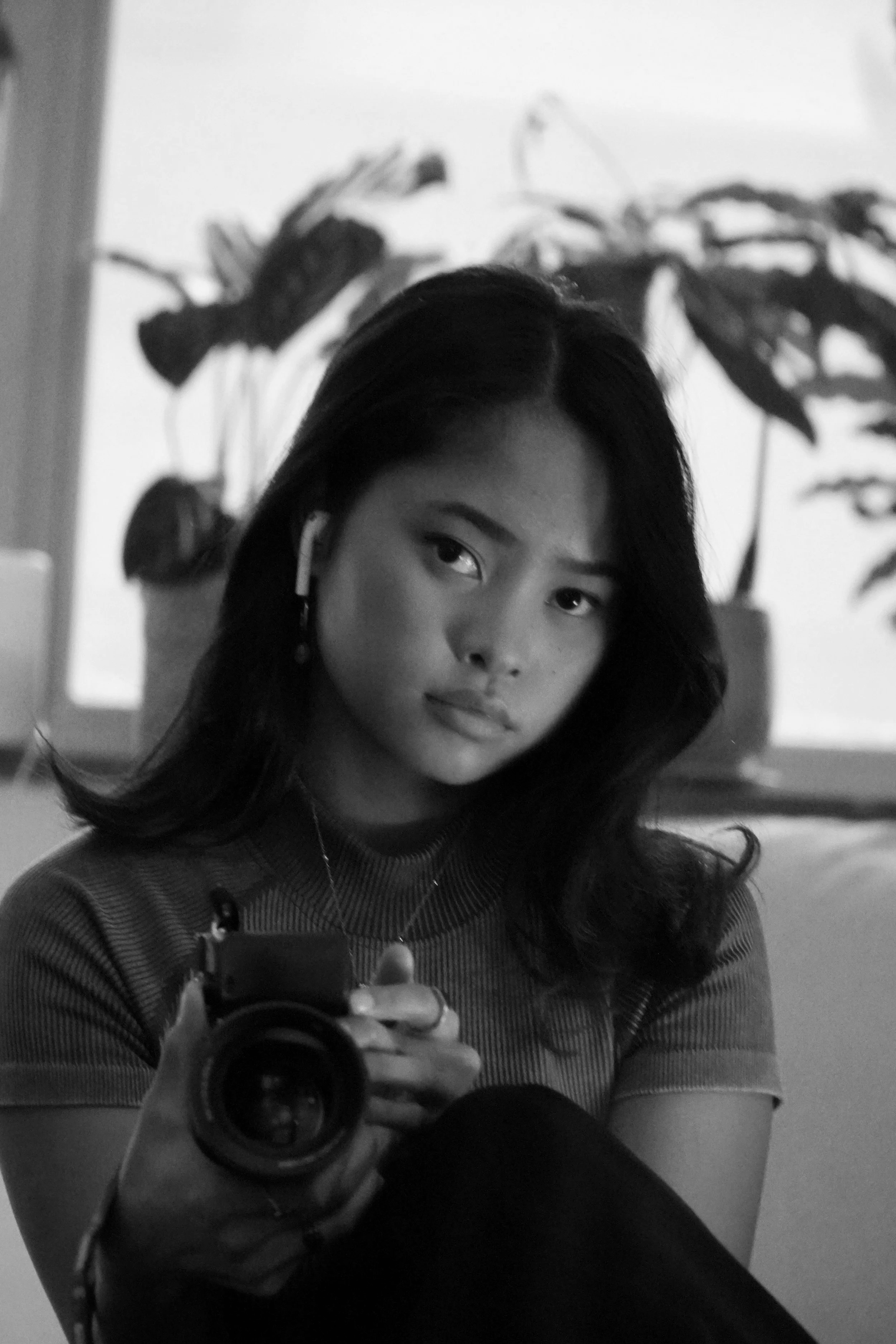 A black and white photo of a woman taking a selfie in front of a mirror, with large leafy plants in the background.