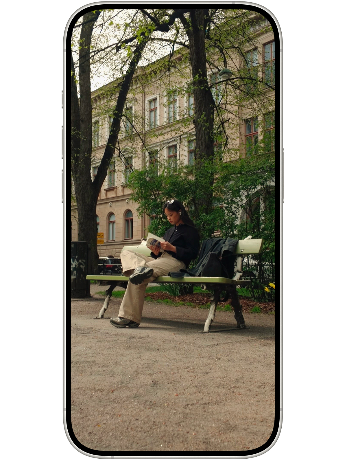 A young woman with long dark hair sitting on a green park bench reading a book. She is wearing a black jacket, beige pants, and gray shoes. There are trees and a large building in the background, and her backpack rests on the bench next to her. The scene is set in an urban park with a dirt pathway.