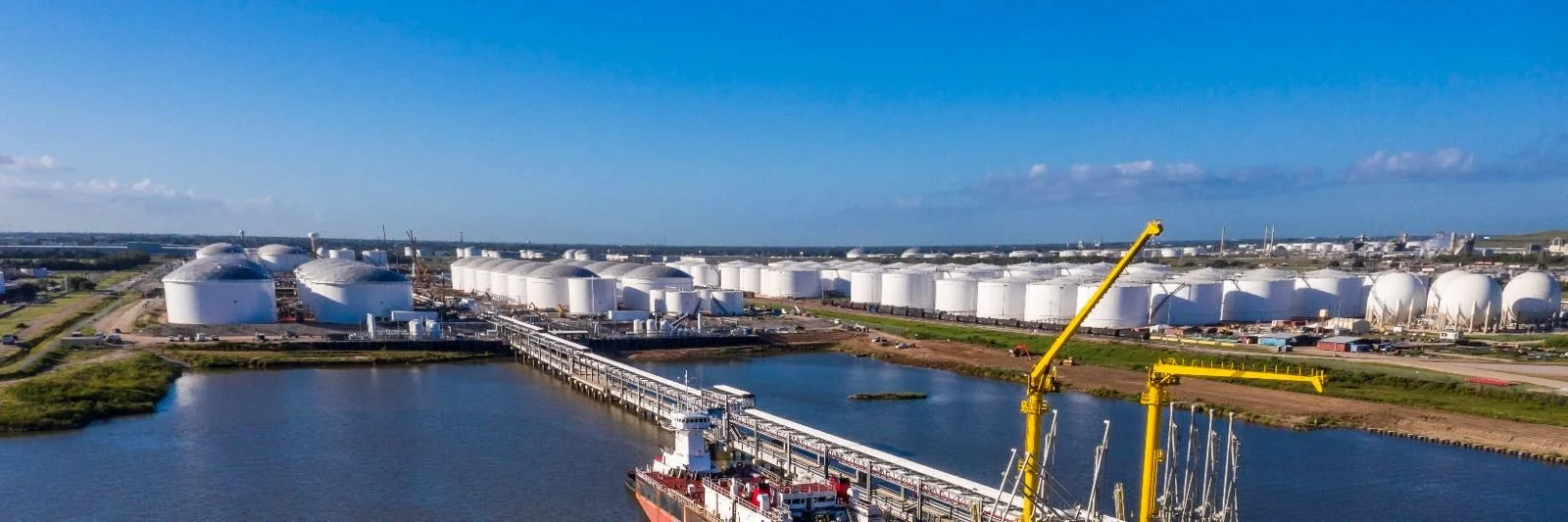 Shell Kazakhstan B.V Aerial view of an industrial port with white storage tanks, a ship docked at the pier, and yellow cranes against a blue sky.