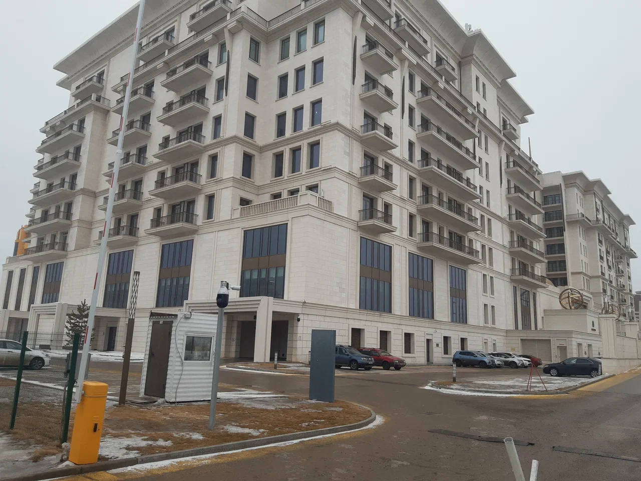 Shell Kazakhstan B.V large multi-story residential building with many balconies and windows. The building is white with some modern architectural details. There are cars parked in the driveway and some snow on the ground.